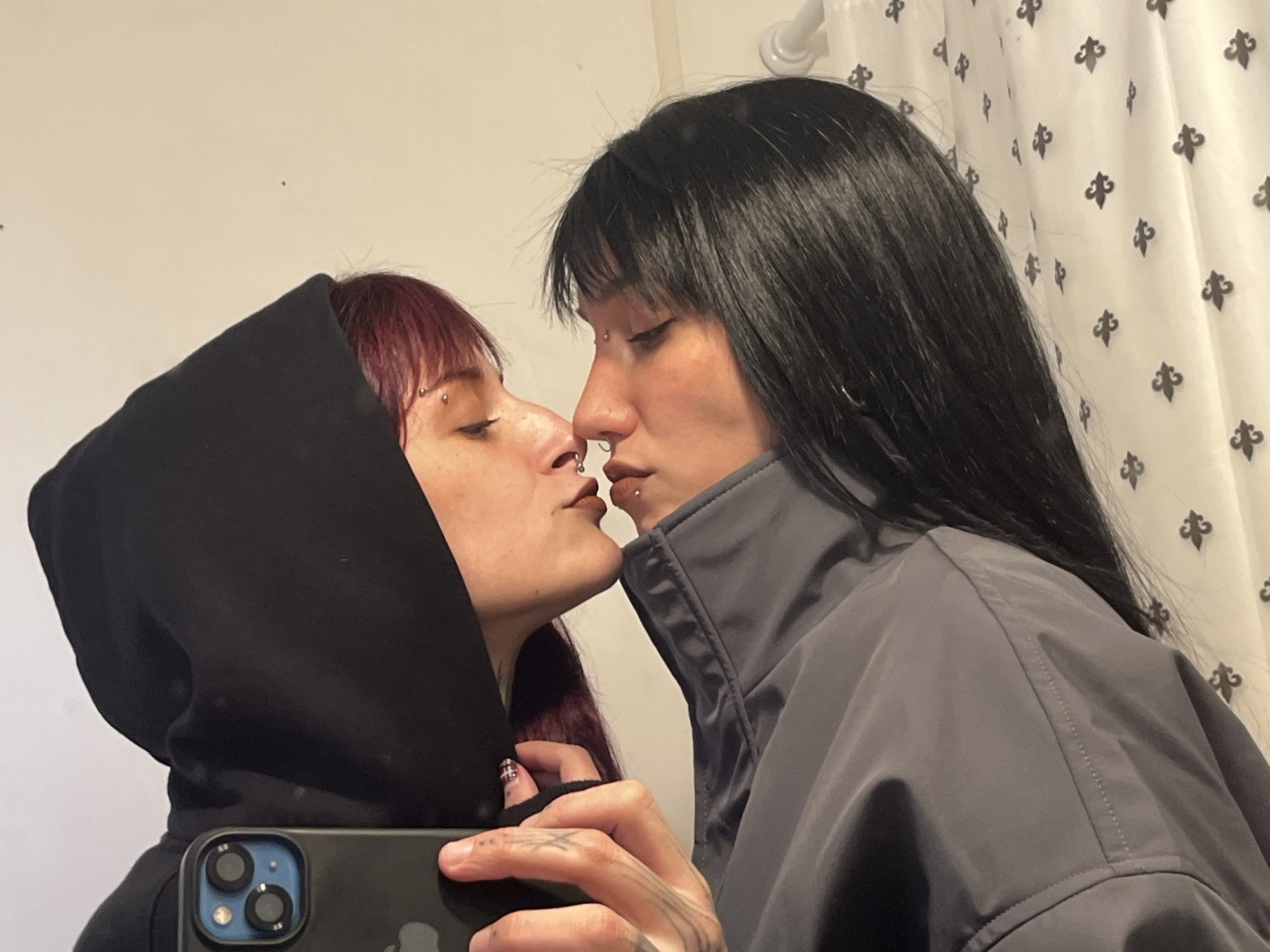 An intimate indoor mirror selfie captures two individuals sharing a moment of affection. The person on the left, with dark red hair, is wearing a black hooded top. They hold a dark-colored smartphone in their right hand, reflected in the mirror as they take the picture. This individual has visible eyebrow and nose piercings, and their eyes are closed as their lips are positioned close to the other person.
The person on the right has long, straight dark hair with bangs and is wearing a grey jacket. They also have multiple facial piercings, including a septum ring and a labret piercing. Their eyes are downcast, almost closed, indicating a tender moment. The two individuals are positioned very closely, with their lips almost or lightly touching, suggesting a kiss or a close embrace.
Notable details include tattoos with intricate line patterns visible on the fingers and back of the hand holding the phone, and one fingernail sports dark polish with a subtle design. The background is simple: a plain white wall on the left and a white curtain adorned with a repeating dark fleur-de-lis pattern on the right. The scene conveys a strong sense of intimacy and connection, taking place in a private, indoor setting, potentially a home in Buenos Aires, Argentina. No explicit text is visible in the image.