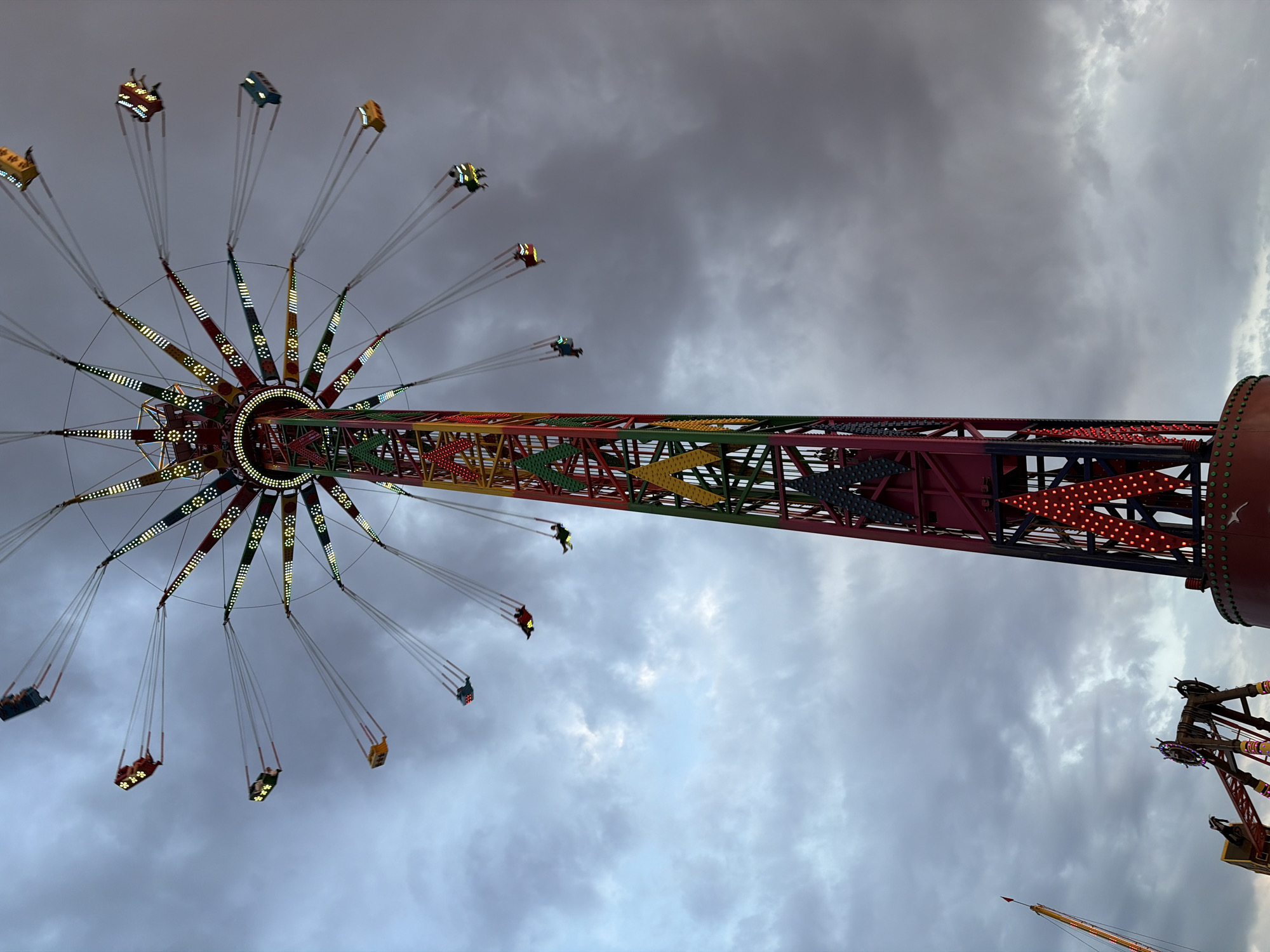 An extreme wide shot captures a brightly lit amusement park ride against a cloudy, twilight sky. The ride, a tall tower with a spinning carousel of swings, dominates the center of the frame. The swings, each carrying one or two passengers, are suspended by cables and appear to be in motion, fanning out from the central tower. The tower itself is a colorful, multi-colored structure with red, green, and yellow accents, illuminated by numerous small, bright lights that form decorative patterns. In the foreground, at the bottom of the image, other amusement park structures are visible, including a partial view of another ride with metal supports and lighting. The overall scene suggests a vibrant and energetic atmosphere, typical of a fair or carnival.