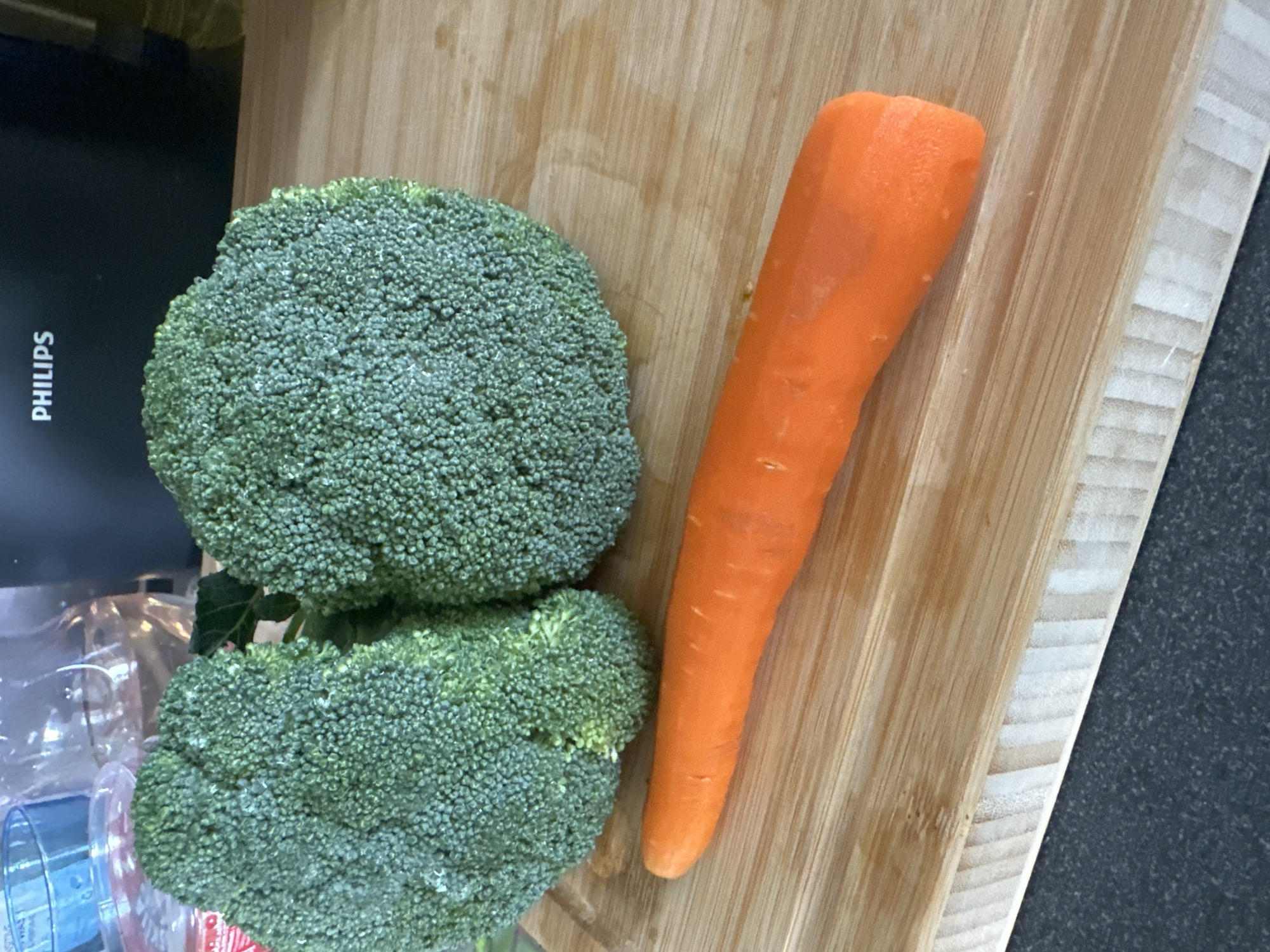 This media file contains an overhead shot of two heads of broccoli and a peeled carrot on a wooden cutting board. The broccoli heads are a vibrant green, with their characteristic florets clearly visible. One head of broccoli is larger and sits on top, while the other is slightly smaller and positioned below it. To the right of the broccoli is a single, peeled carrot, its bright orange color contrasting with the green of the broccoli and the light brown of the cutting board. The cutting board itself has a natural wood grain pattern. In the background, to the left of the cutting board, a dark, reflective surface with the white text "PHILIPS" vertically oriented is visible. There are also some clear plastic containers or wrappers in the lower left corner. The scene appears to be a kitchen or food preparation area, with the lighting suggesting it could be daytime or well-lit indoor lighting. There are no people present, and the focus is solely on the fresh produce.