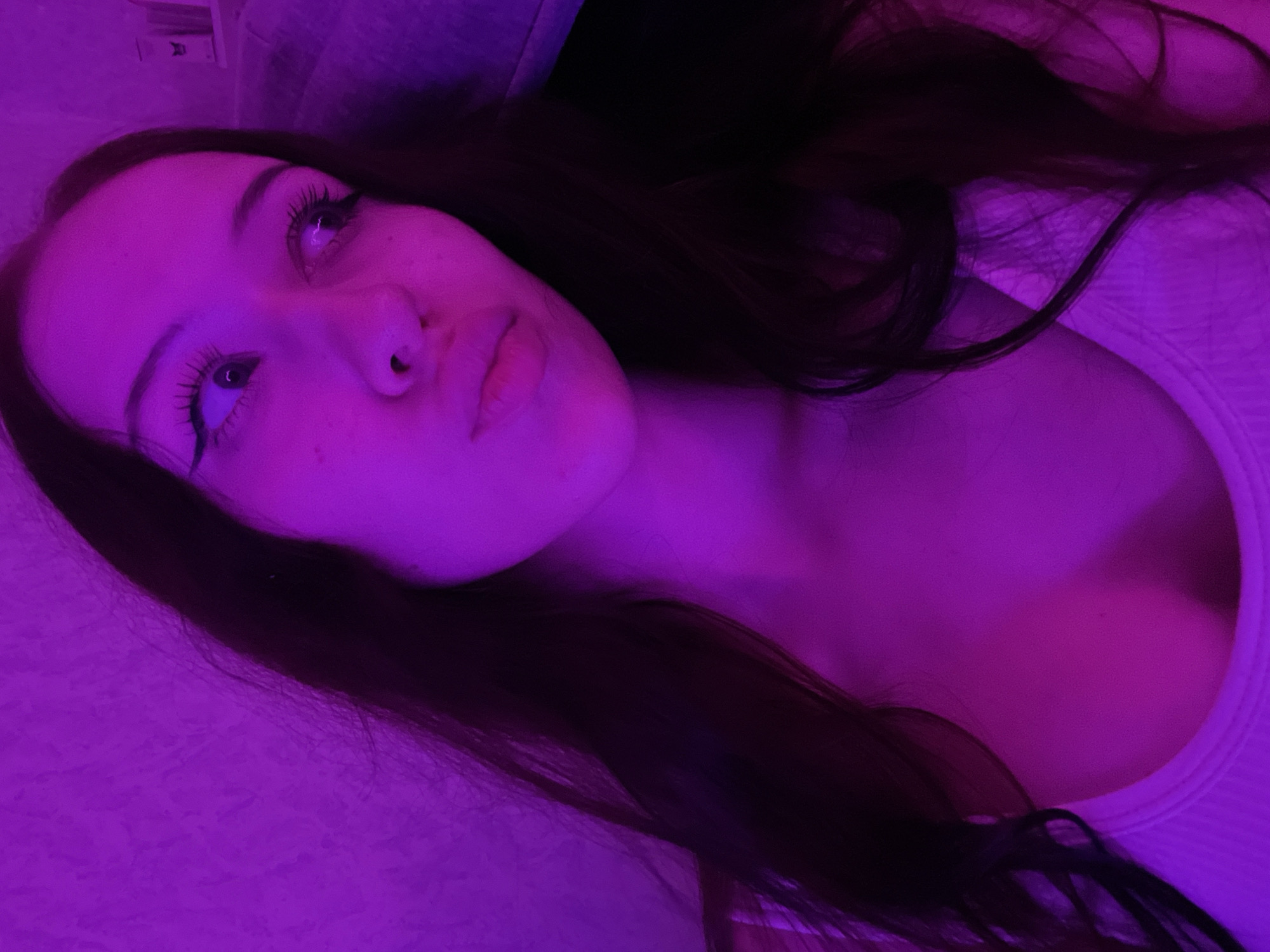 The image is a close-up selfie of a young woman illuminated by purple light. She has long, dark hair that falls around her face and shoulders. Her eyes are looking away from the camera, and her lips are slightly parted. She is wearing a white tank top. The background appears to be a wall with a textured surface, also lit by the purple light. There is a shelf with some objects visible in the upper right corner of the image, though they are indistinct due to the lighting and resolution. The image is a portrait, focusing on the woman's face and upper body. There is no indication of the city of Votkinsk, Russia, or any specific location within it.