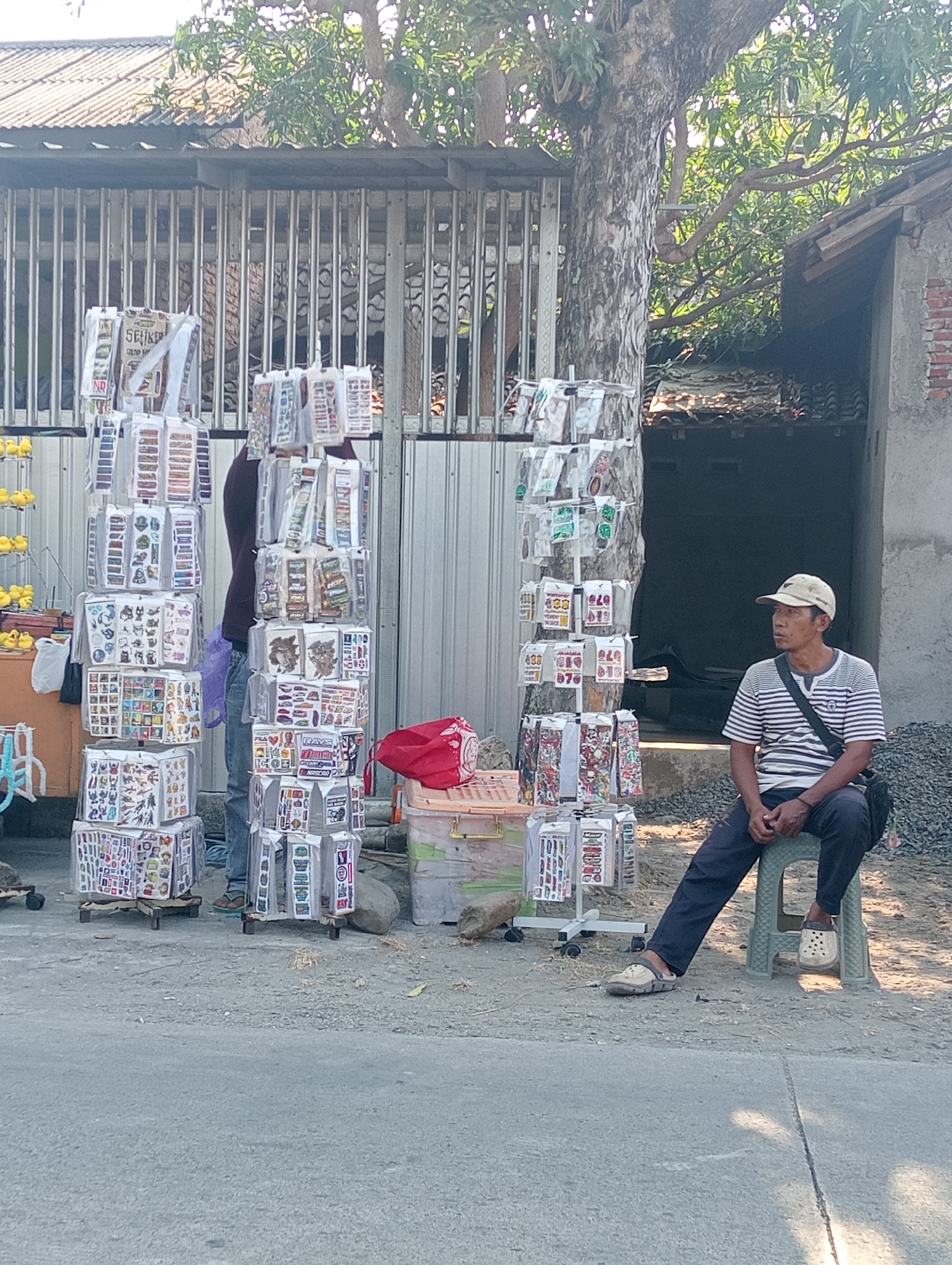 A man wearing a striped t-shirt and dark pants sits on a green plastic stool in front of a stall selling stickers. He wears a beige baseball cap and a black bag is slung across his body. The stall is set up on a paved street in Tanjunganyar, Indonesia, with a metal fence and a building in the background. Several tiered racks are filled with various packs of stickers, some displaying cartoon characters and others with text. A large tree with green foliage is visible behind the stall, and debris, including rocks and soil, is scattered around the area. To the left of the stall, a smaller orange cart with yellow objects is partially visible.