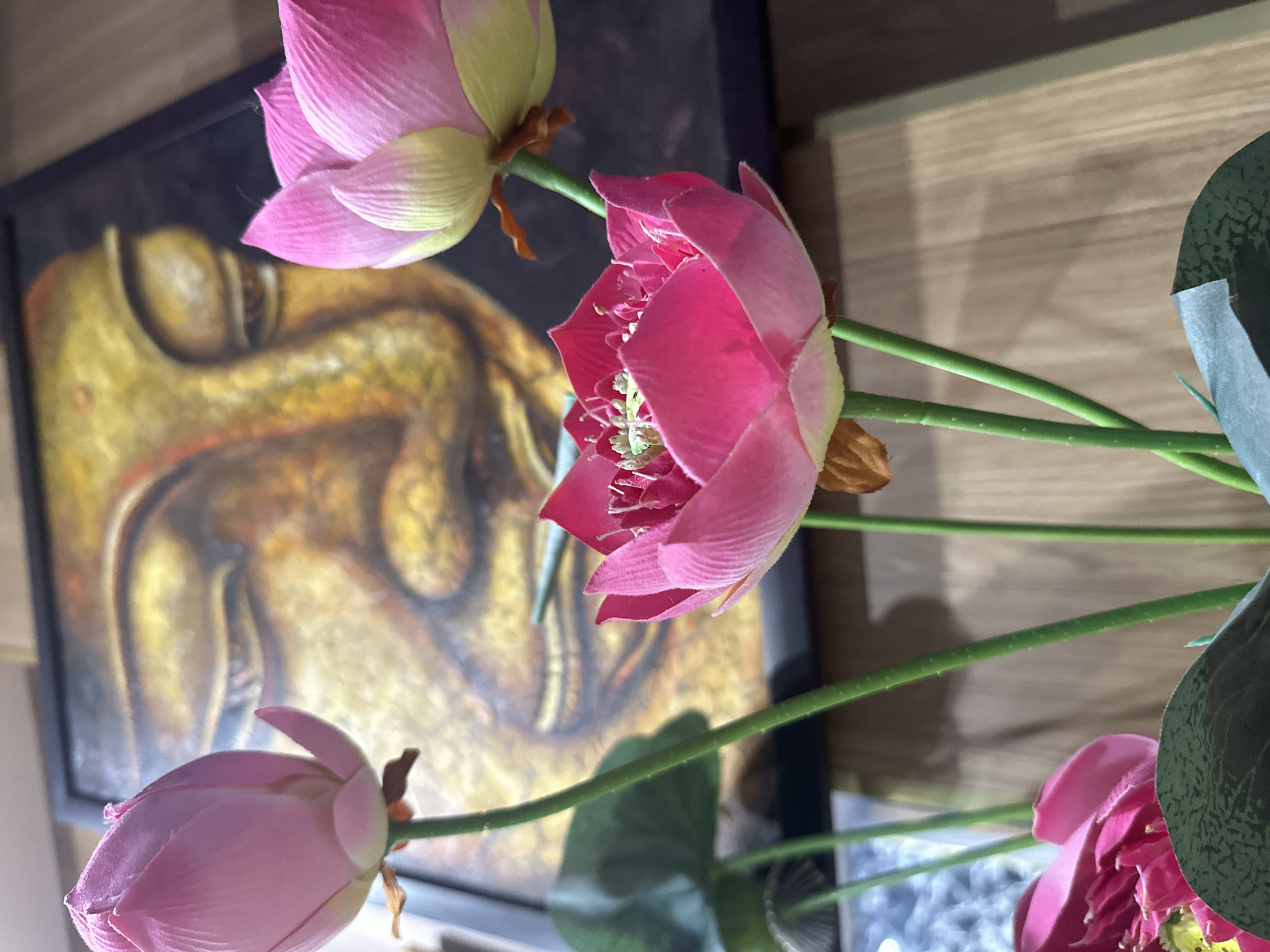 A close-up shot captures artificial pink lotus flowers in partial bloom, set against a background of a framed image depicting a golden figure, possibly Buddha. The scene includes a blurred, wood-grain surface.