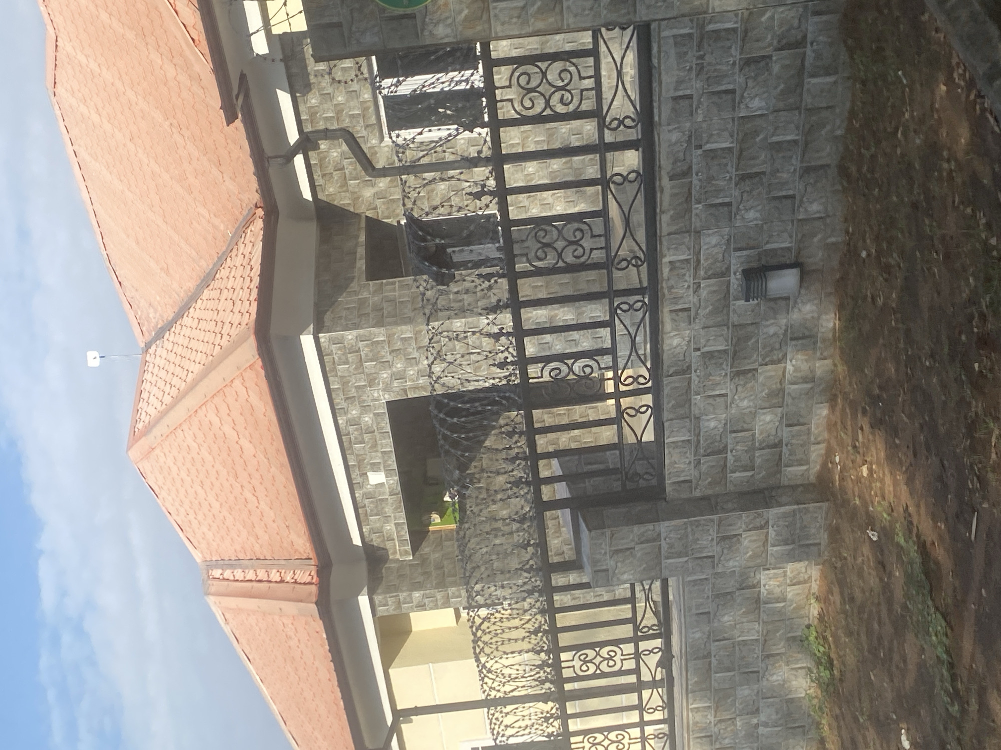 The image shows a security gate in front of a building with a red tile roof. The gate is made of black metal and has a decorative design. There is barbed wire wrapped around the gate. The building is made of grey stone. The location is Unknown, Unknown. The sky is visible in the background.