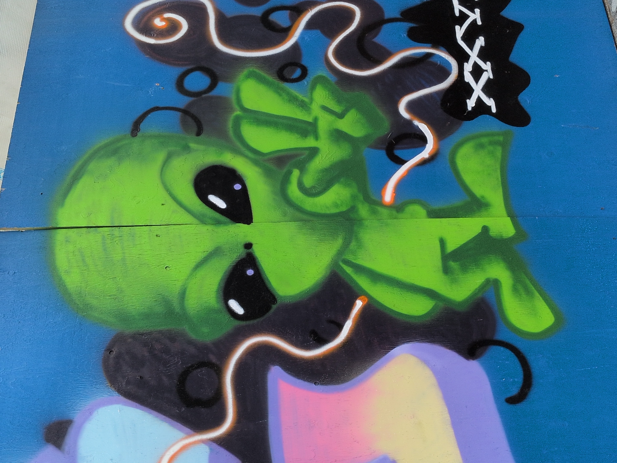 This is a graffiti image on a blue wooden surface. The central subject is a cartoonish green alien with large black eyes, one of which has a purple pupil. The alien is depicted in a split style, with its left side slightly darker and less detailed than its right side. The alien's right hand is raised in a peace sign. To the left of the alien, there are abstract shapes in purple and light blue, with a pink and yellow gradient. Swirling white and orange lines are spray-painted around the alien, with black accents and circular shapes. To the lower right of the alien, there is a black jagged shape with white "XXX" written on it, resembling stitching. The graffiti is located in Helsinki, Finland.