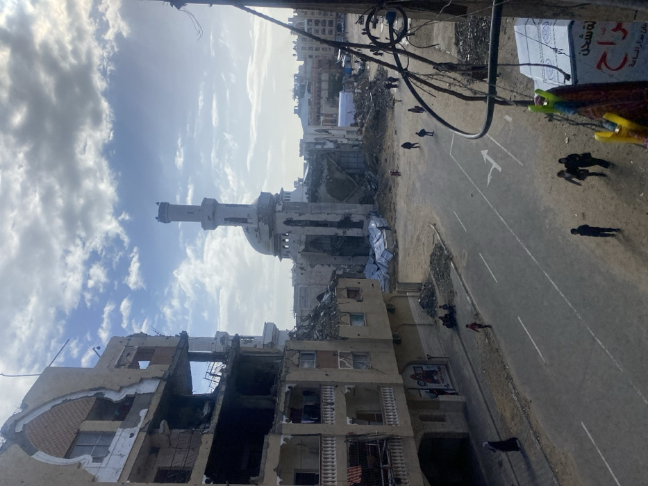 The image displays a street in Khan Yunis, Palestinian Territory, showing extensive structural damage under a partly cloudy sky. On the left, a multi-story building exhibits severe damage, with multiple floors partially collapsed, exposed interiors, and missing sections of its facade and roof. Further into the scene, a large mosque with a prominent minaret and dome is visible, also heavily damaged, with sections of its walls missing and rubble surrounding its base. Blue tarpaulins cover portions of the debris field near the mosque.
A paved road with a white double-line marking and a right-turn arrow runs through the center of the frame. The areas immediately adjacent to the road are composed of dirt and piles of rubble. Multiple individuals are present: approximately six adults and four children are walking on the road, while two adults and three children are gathered on the sidewalk to the left, near the base of the damaged building. One woman in a dark abaya walks on the far left sidewalk. In the foreground right, damaged electrical wires or cables hang loosely. A partial view of a shop sign with Arabic script and some clothes pegs hanging from a railing are visible in the extreme right foreground.