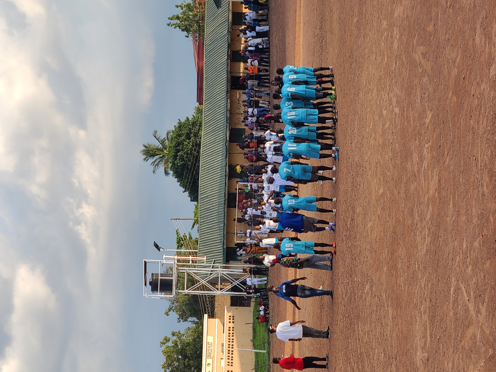 An outdoor scene captures a group of individuals on a reddish-brown dirt field under an overcast sky. In the foreground, approximately 11 men, identified as a team, stand in light blue jerseys with numbers (e.g., 7, 11, 20, 18, 3, 6, 9, 2, 13) on their backs, facing away from the camera. They are arranged in a loose semi-circle. To their left, several other men are present: one in a red shirt on the far left, another in a white shirt and gray pants, one in a dark blue shirt labeled "OFFICIAL" facing the team, and a man in a multi-colored patterned shirt.
In the mid-ground, a crowd of spectators, numbering over 50, lines the area near several buildings. One prominent building is light beige with "SUWASH 2023 PROJECT" and "MALE TOILETS" inscribed on its facade. Adjacent to it is a structure supporting a black water tank, featuring a solar-powered street light. Further right is a building with a green corrugated roof, and faint lettering on its wall reads "THIS IS ASSEMB". A goalpost structure is partially visible near the green-roofed building, suggesting a sports field. Various trees, including a palm tree, are scattered throughout the background. Electrical poles and wires are also visible. The setting is likely a community or school ground in the city of Unknown, Unknown.
