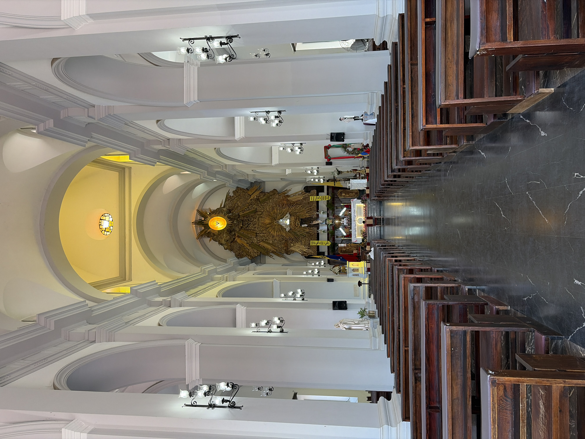 The image depicts the interior of the Ciudad Bolivar Cathedral, located in Ciudad Bolivar, Venezuela. It is a wide-angle, high-angle, centered shot showing the nave of the church. The architectural style includes a series of arches supported by white pillars. Black, marble-like flooring reflects light. Dark wooden pews line both sides of the central aisle. Ornate chandeliers illuminate the space. At the far end of the nave is the altar area, adorned with a large, golden, sunburst-like structure. Distant figures can be seen around the altar. The overall ambiance is one of grandeur and historical religious significance.