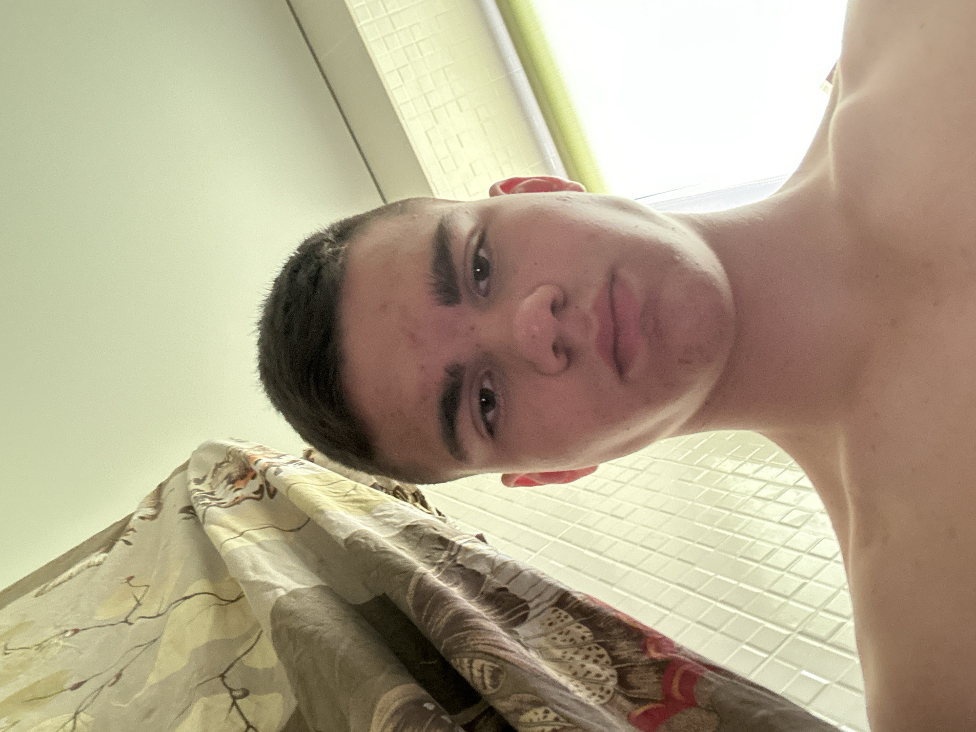 The image is a selfie taken from a low angle, looking up at a young male. He has short, dark hair and appears to be in a bathroom, indicated by the tiled wall and a shower curtain visible on the left. The lighting suggests it might be daytime, with light coming from a window on the right. The background includes a patterned shower curtain with floral motifs and a tiled wall with small, square tiles. The man is shirtless, and his expression is neutral. The image is of moderate quality with some noise.