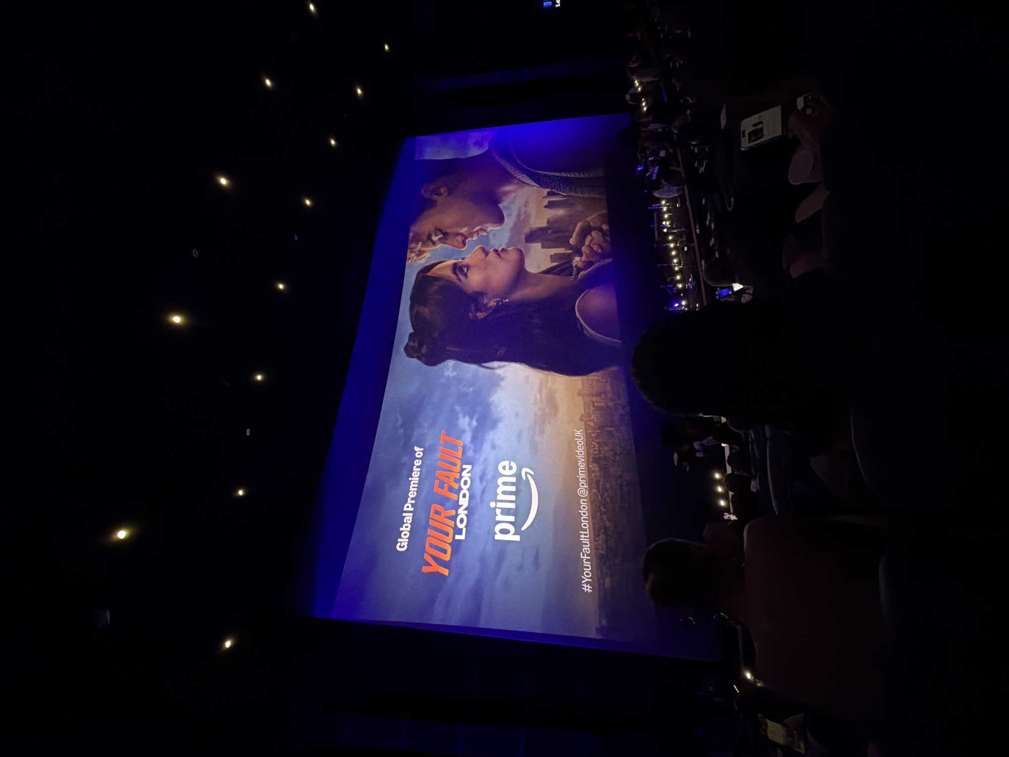 The image depicts a global premiere event for the show "Your Fault London" at a cinema in London, United Kingdom. The large screen displays the show's title in bold orange letters, along with the Amazon Prime Video logo. A promotional image of a man and a woman gazing at each other is also visible. The cinema hall is dimly lit, with rows of seats occupied by an audience. A few individuals are visible in the foreground, facing away from the camera, looking towards the screen. The ceiling of the hall has multiple circular light fixtures. The overall scene suggests a premiere screening, likely before the show's official release.