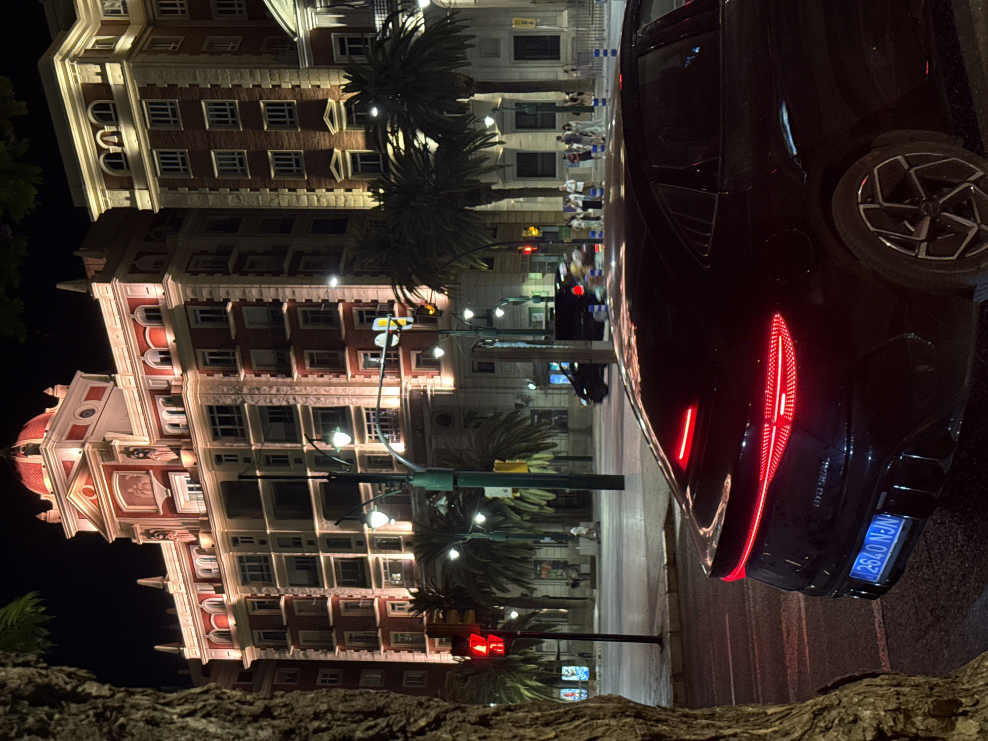 A black car is parked on a city street at night, with its brake lights illuminated. The car has a license plate that reads "2640 N-JN". In the background, there are ornate buildings with red and cream facades, illuminated by warm light. Palm trees are visible along the street. A traffic light with a red signal is also present. A few people are standing on the sidewalk in the distance. The scene appears to be in Málaga, Spain, given the architecture and palm trees.