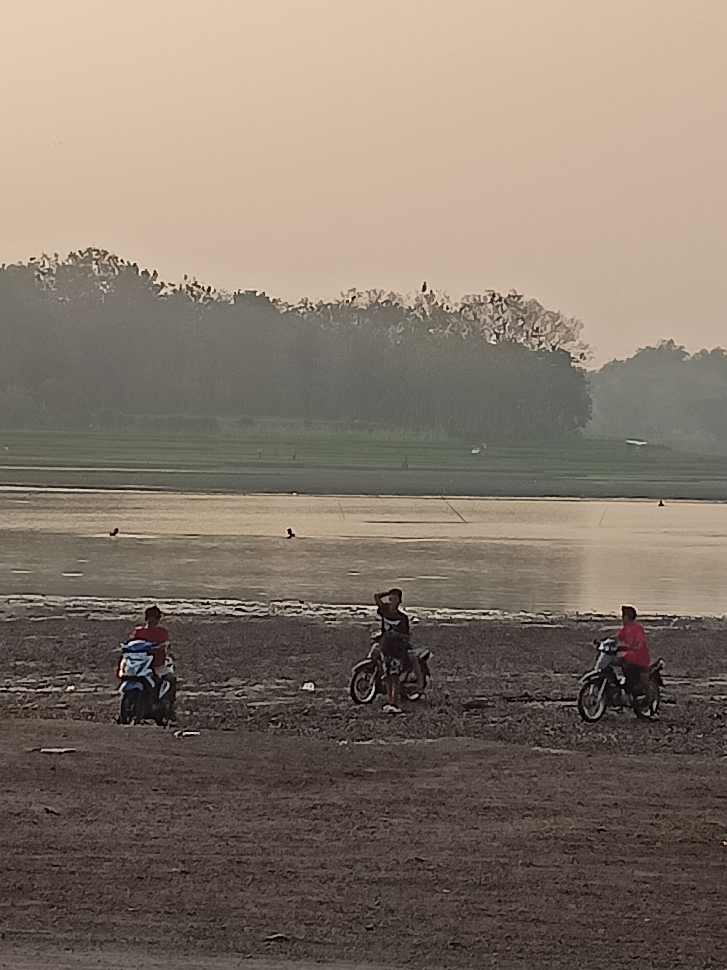 A hazy, low-angle photograph depicts three individuals on motorcycles at the edge of a body of water, likely a lake or river, in Sambiduwur, Indonesia. The foreground is dominated by a dry, barren shoreline covered in small pebbles and dirt. In the middle ground, the water reflects the muted, pale sky, and some indistinct objects, possibly birds or debris, are visible on its surface. Beyond the water, a green field stretches out, backed by a dense line of trees shrouded in mist.
On the shoreline, from left to right:
1. A person wearing a red shirt is seated on a blue and white motorcycle.
2. A person wearing a black shirt is sitting on a motorcycle, with their right hand raised to their head.
3. A person wearing a red shirt is riding a motorcycle.
The overall atmosphere is serene and somewhat distant due to the haze and low resolution of the image.