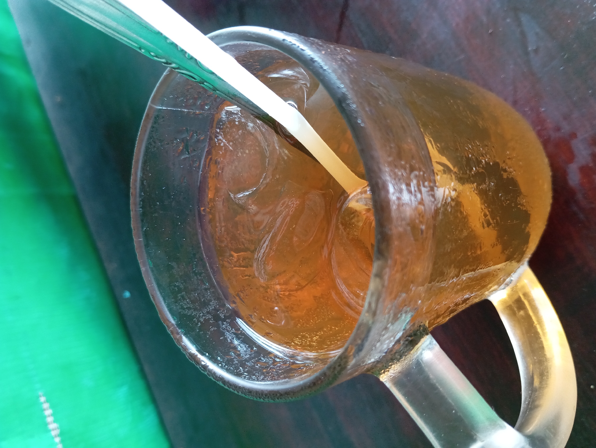 A glass mug filled with iced tea and a green and white straw sits on a dark wooden table. In the background, a green surface is visible. The scene is located in Lemahbang, Indonesia.