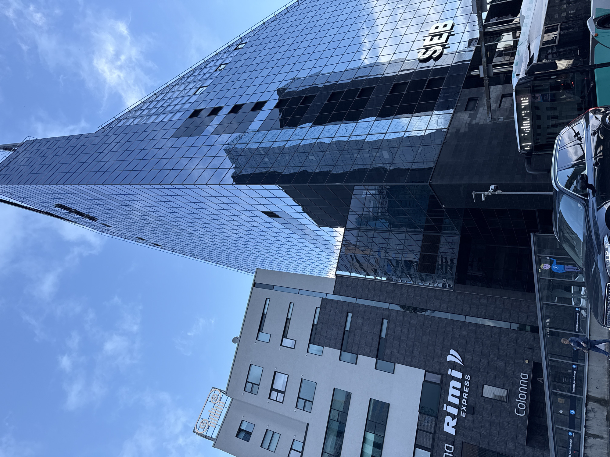 In Tallinn, Estonia, an urban scene unfolds with modern architecture under a partly cloudy sky. Dominating the frame is a tall, sleek skyscraper clad in reflective blue glass, its facade mirroring the sky and surrounding buildings. To its left, a contrasting building features a lighter, textured gray exterior with numerous rectangular windows of varying sizes. A prominent sign on this building reads "Rimi EXPRESS" with a stylized "V" logo and below it, "Colonna". Further to the right, the glass skyscraper displays a prominent illuminated sign spelling out "SEB". In the foreground, a dark-colored SUV is parked, and a bus with its digital display lit up is visible to the right of the SUV, partially obscuring the lower portion of the SEB building. A few individuals are present, one walking in the mid-ground on a sidewalk near the Colonna sign, and another, a cartoon-like figure of a person with a blue "P" symbol, is positioned near the bus stop area. The street level shows a pedestrian walkway with signage related to parking. The overall impression is one of a contemporary commercial district.