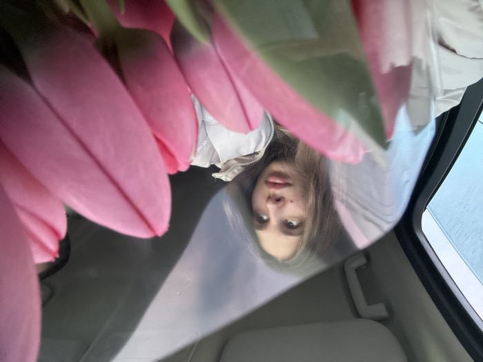 The image depicts the interior of a vehicle in Slavyansk-na-Kubani, Russia. A young female individual is situated in the left-center of the frame, facing slightly rightward with her gaze directed upwards. She has long, light brown hair and is wearing a light-colored, possibly white or beige, quilted jacket. A small dark mark or mole is visible on her right cheek. In the foreground, occupying the right half of the image, is a bouquet of multiple pink tulip buds, tightly closed and displaying some green stem and leaf elements. The tulips appear to be wrapped in a transparent material, causing reflections across their surface. The vehicle's interior shows a light-colored grab handle and part of the headliner in the upper sections. A dark vertical element, consistent with a car's pillar or door frame, is present on the far left. Through a window in the upper-left, a blurred outdoor environment with blue and grey tones is visible, suggesting an overcast sky. The scene is illuminated by ambient daylight.