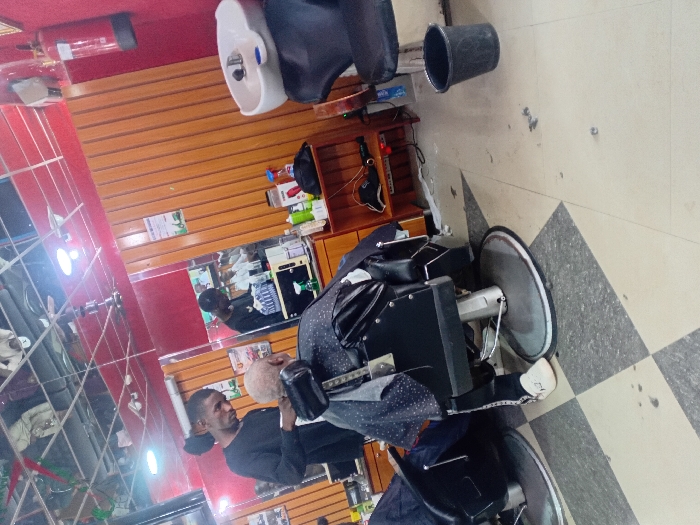 The scene is set inside a barbershop or hair salon in Dorayi, Nigeria. A male barber, wearing a black long-sleeved shirt, stands behind a seated client, actively trimming the client's short, light-colored hair with clippers. The client is draped in a black and white polka-dotted cutting cape and occupies a black barber chair with a chrome base. The barbershop interior features red-painted walls with lower sections of vertical wooden paneling. The ceiling consists of reflective panels, from which light fixtures and a ceiling fan are suspended. The floor is tiled in a checkerboard pattern of light grey and dark grey squares. To the right, a white ceramic hair wash basin is mounted, accompanied by a black chair, and a black plastic bucket rests on the floor beneath it. A red fire extinguisher is mounted on the red wall in the upper right. Mirrors cover a significant portion of the back wall, reflecting the barber, the interior, and a television screen displaying what appears to be individuals. Various hair care products and tools are arranged on a wooden shelving unit and countertop behind the barber and client. A second black barber chair is partially visible on the far left.