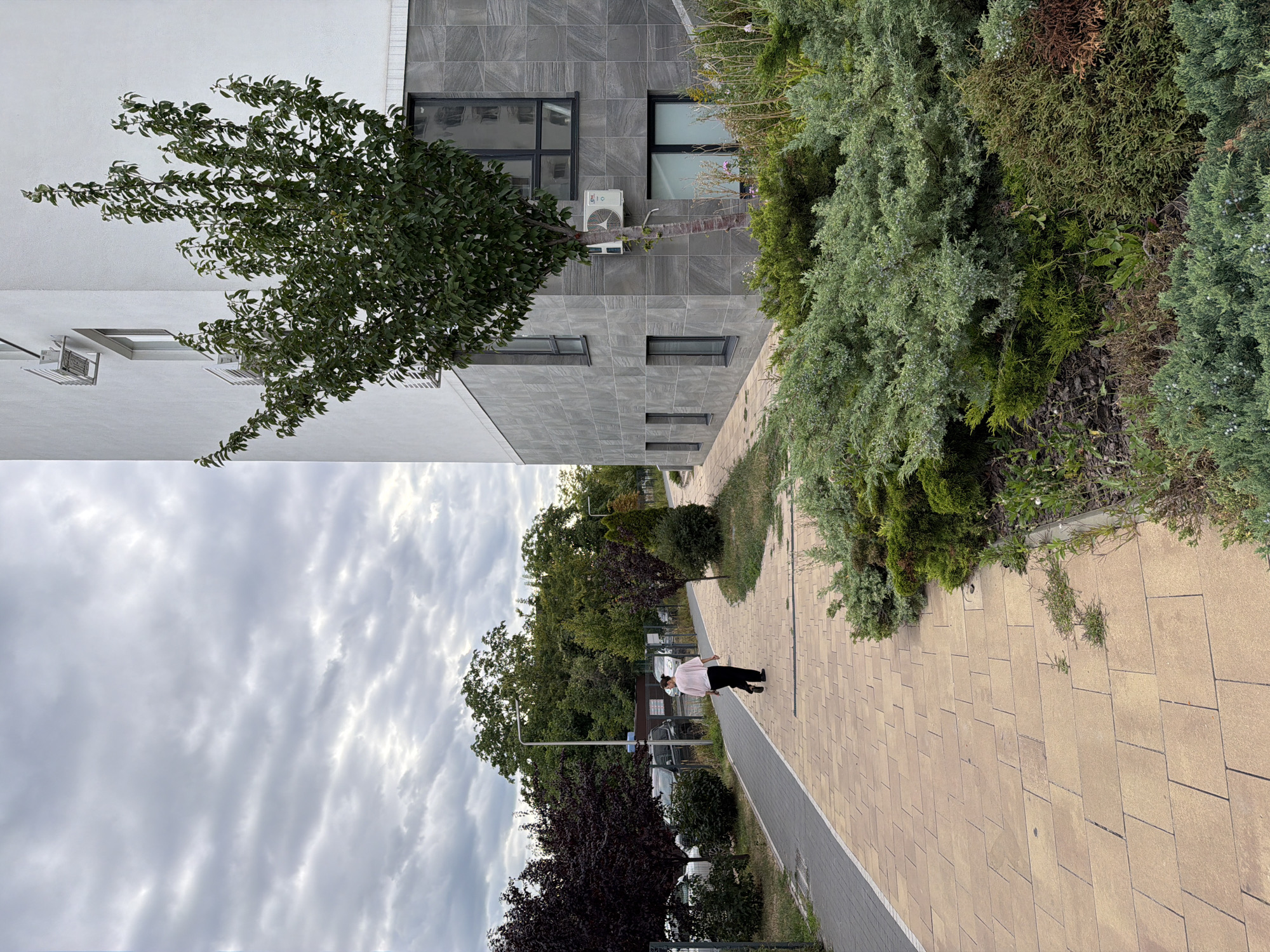 The image shows a pedestrian walkway in front of a modern building in Kyiv, Ukraine. A person is walking away from the camera on the walkway, dressed in a pink shirt and black pants. The building has a facade of grey stone tiles and white upper levels, with several windows and an air conditioning unit visible. A young tree stands in front of the building, near the walkway. The walkway is paved with light brown rectangular tiles. To the right of the walkway is a landscaping area with various green shrubs and plants. In the background, there are other trees, parked cars, and streetlights. The sky is overcast with grey clouds.