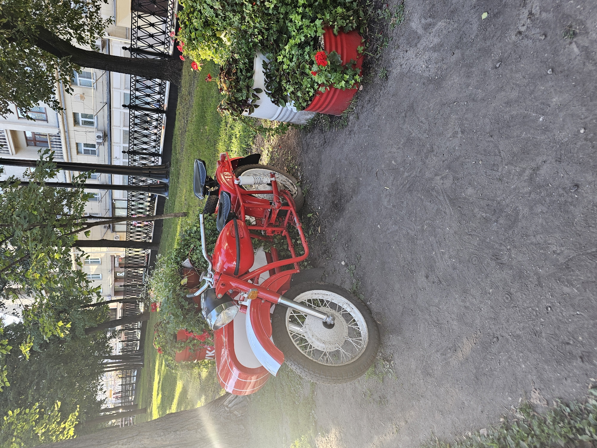 A red motorcycle with a sidecar is parked on a dirt path next to a grassy area in Moscow, Russia. The motorcycle is vintage and appears to be in good condition. The sidecar is also red and white. The grassy area is decorated with potted plants, including red geraniums. In the background, there is a building with several windows and a wrought-iron fence. The sun is shining, casting long shadows. The ground is a mixture of dirt and grass, with some fallen leaves.