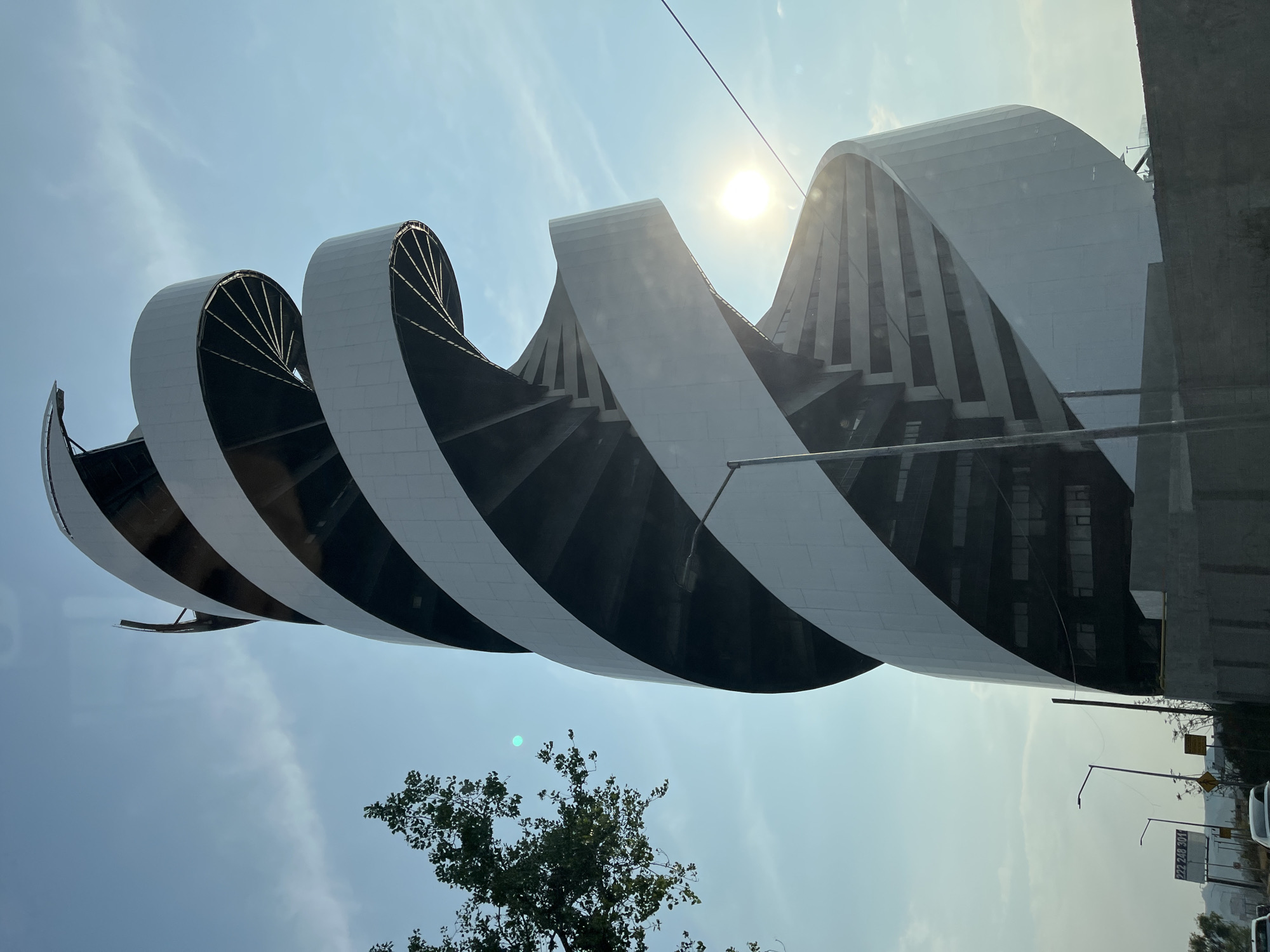The image captures a modern architectural structure in San Bernardino Tlaxcalancingo, Mexico. The building features a distinctive, undulating design with alternating white and dark surfaces, creating a wave-like effect. The sun is positioned behind the building, casting a bright glow. A tree with green foliage is visible in the lower portion of the image. Streetlights, signage, and vehicles are present, suggesting an urban or suburban environment. The overall atmosphere is bright and clear.