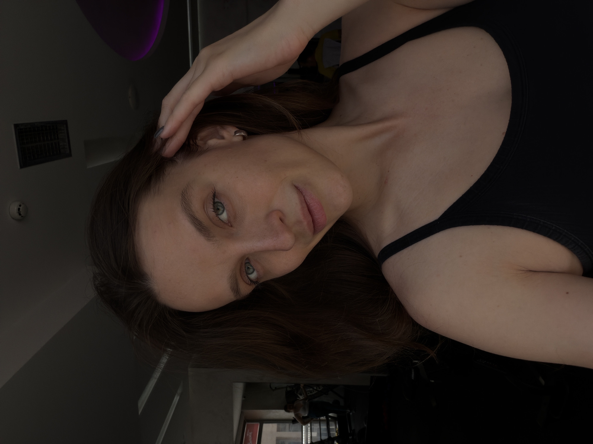 A young woman with long brown hair and green eyes is posing in a dimly lit room. She is wearing a black tank top. Her left hand rests on her head. In the background, through a window, there appears to be a glimpse of an interior possibly in Budva, Montenegro. There are indistinct figures in the background, seemingly engaged in activity, perhaps in a gym or fitness center.