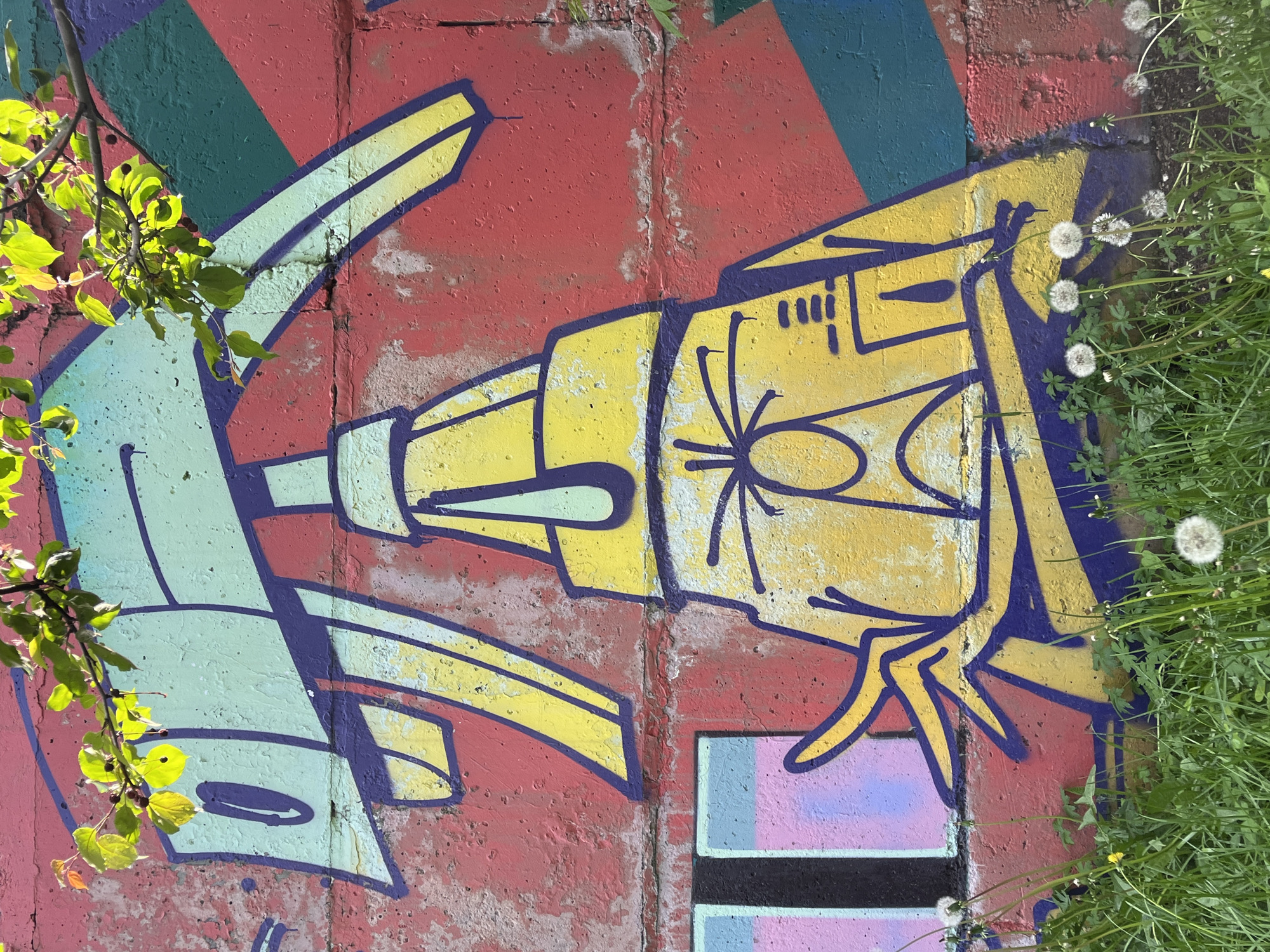 The image shows a graffiti artwork on a red brick wall, depicting a yellow spray bottle with a face and arms. The spray bottle has a blue outline and a light blue nozzle. Green branches with leaves hang from the top of the frame, partially obscuring the graffiti. The wall is covered in a red and pink abstract pattern with green and blue diagonal lines. In the foreground, green grass and dandelions are visible. The graffiti is located in the city of Krasnoyarsk, Russia.