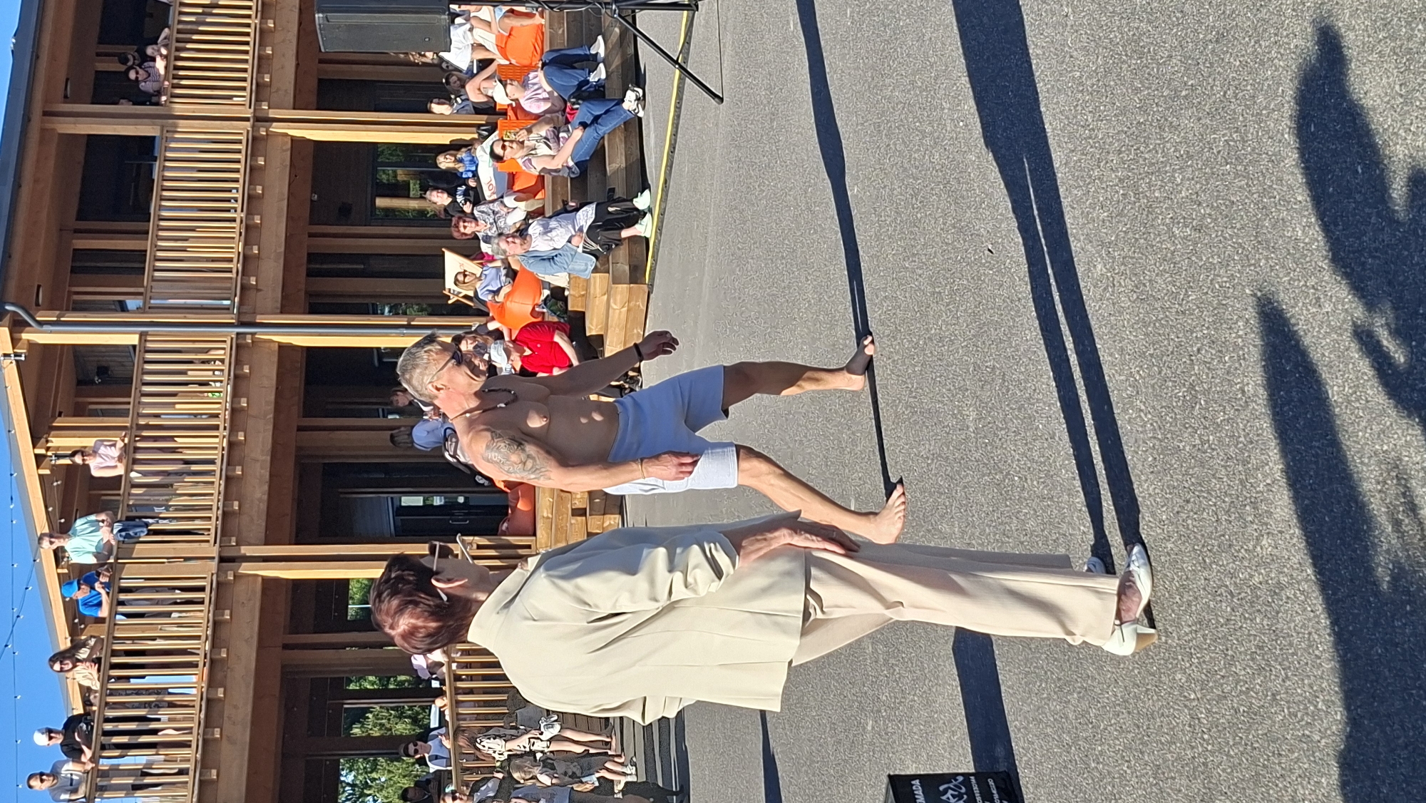 In the city of Tallinn, Estonia, a man with tattoos on his arm and shoulder walks barefoot across a paved area in front of a wooden building with multiple levels and balconies. He is wearing light blue shorts. To his left, a woman wearing a beige suit jacket and matching pants walks away from the camera, her back to the viewer. She is wearing white heeled shoes. In the background, several people are seated on orange beanbag chairs and wooden steps. The building appears to be a modern structure with large windows and wooden railings. The scene is set outdoors during the day, with sunlight casting long shadows on the ground.