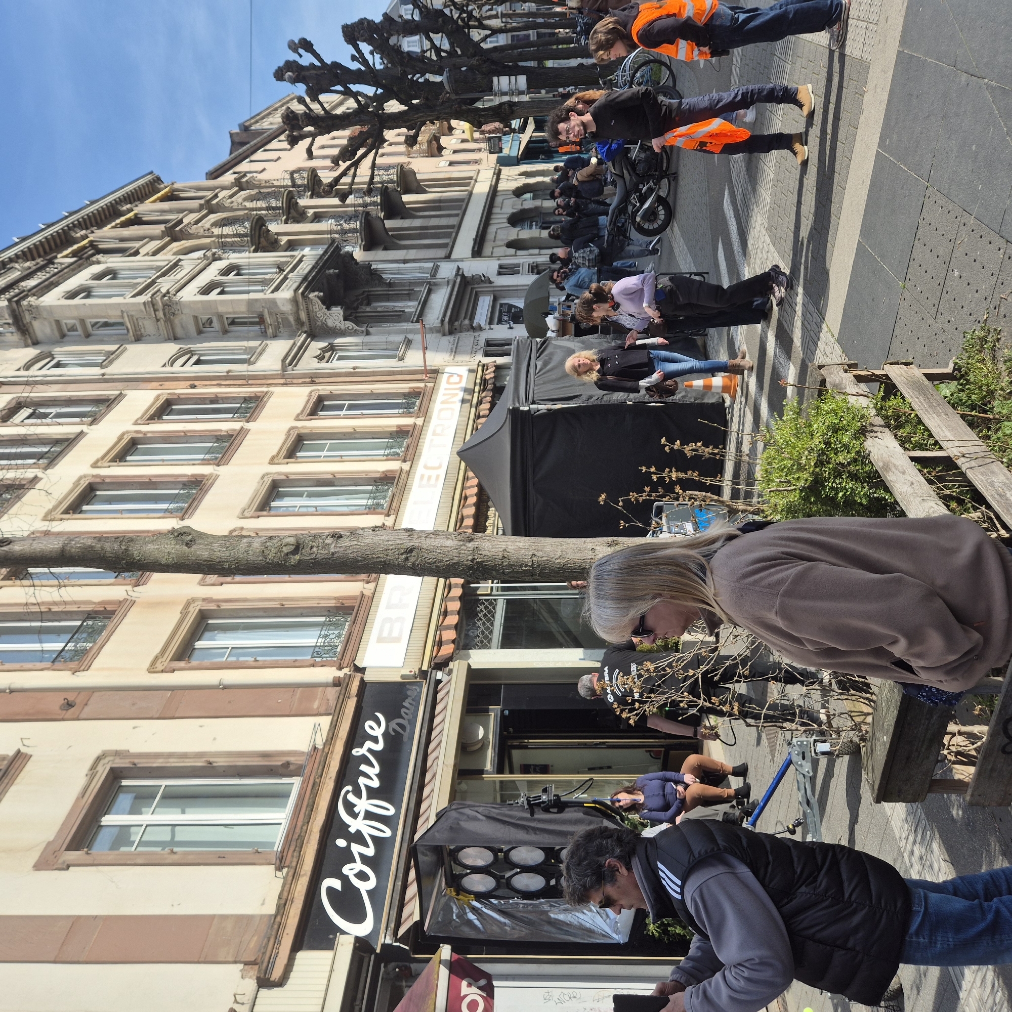 The scene depicts a daytime street view on Rue des Hallebardes in Strasbourg, France, under a clear blue sky, indicating a film or photo shoot in progress.
In the immediate foreground, to the left, a male in a grey sweatshirt and black puffer vest with white shoulder stripes, wearing sunglasses, holds a dark smartphone or camera to capture an image. To the right, a blonde female in a tan fleece jacket and sunglasses stands with hands in pockets, observing the scene. A wooden planter with small green shrubs and bare branches is situated between them.
Mid-ground left features the entrance of a shop signed "Coiffure Dames." A large film lighting fixture with eight circular lights on a stand is positioned on the sidewalk, partially obscured by a black fabric reflector. A blue camera dolly rests nearby. A female in a blue top and brown pants sits on a stool near the shop entrance, while a male crew member in a black "ALTERA" t-shirt walks away.
Further right in the mid-ground, a prominent bare tree trunk stands. Behind it, two black pop-up tents are erected on the sidewalk. A group of individuals are gathered on the sidewalk and street, including a female in a dark coat and blue jeans, another female in a lavender top with headphones around her neck, and a male holding an orange safety vest. Another individual wears an orange safety vest on the far right. A motorcycle is parked on the street. Orange traffic cones are visible on the sidewalk.
The background features multi-story European-style buildings. The building on the left, housing "Coiffure Dames," has a light facade with reddish-brown vertical stripes. Adjacent buildings exhibit light-colored facades; one partially displays "ELECTRONIC," and another further down features ornate grey stone architecture with multiple balconies and decorative carvings. A line of pollarded trees with bare branches extends along the right side of the street. The street is paved and includes a crosswalk. Numerous individuals are visible further down the street.