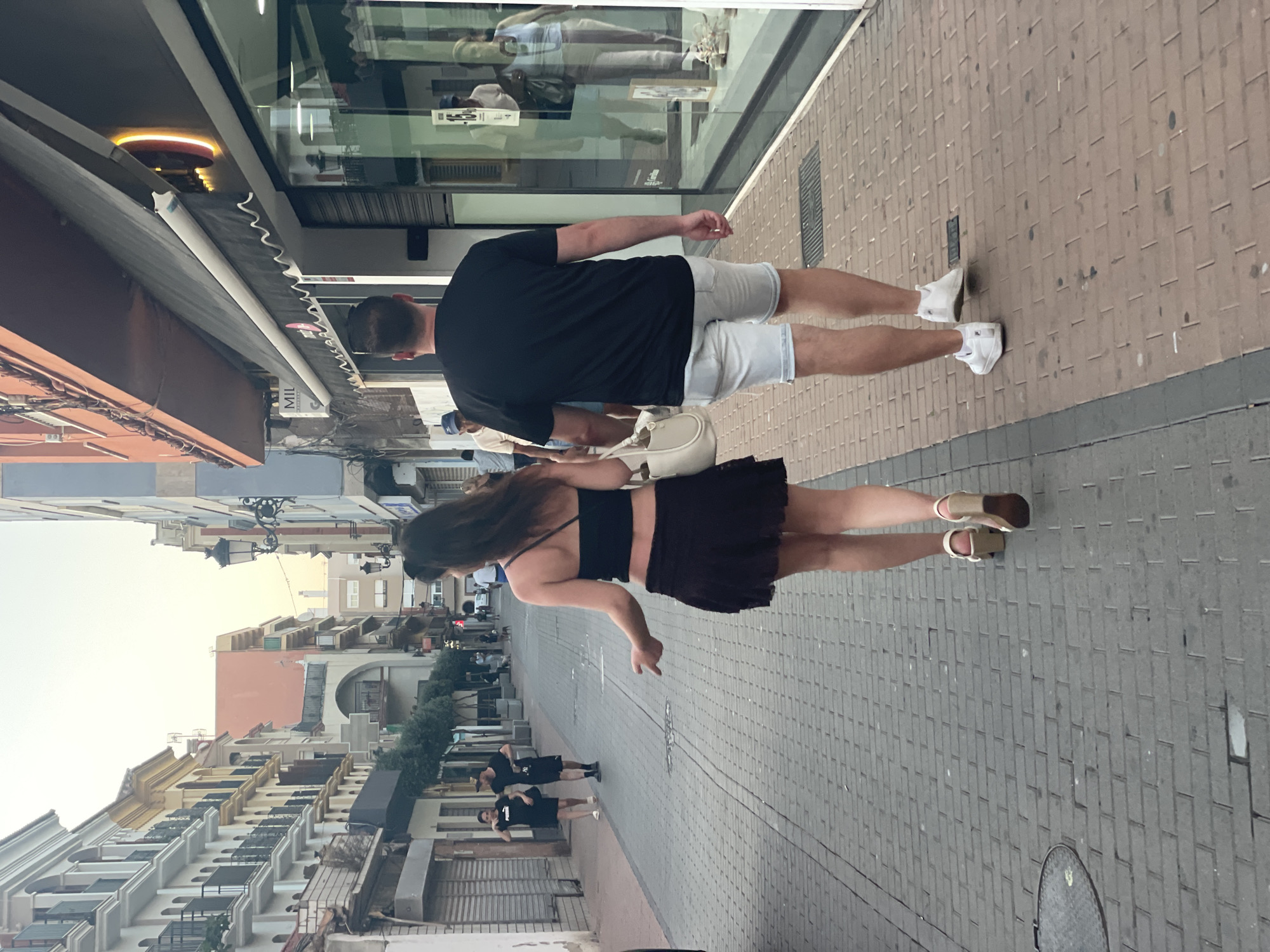In La Línea De La Concepción, Spain, a couple walks away from the camera on a brick-paved street. The woman, on the left, wears a dark crop top and a dark, short, ruffled skirt, with light-colored platform sandals. She carries a light-colored shoulder bag and has dark hair. The man, on the right, wears a black t-shirt and light-colored denim shorts with white sneakers. They are walking on the sidewalk adjacent to shops. The street features buildings with balconies on the left and shopfronts with awnings on the right. Several other pedestrians are visible in the background, as well as trees lining the sidewalk. The sky is overcast and light.