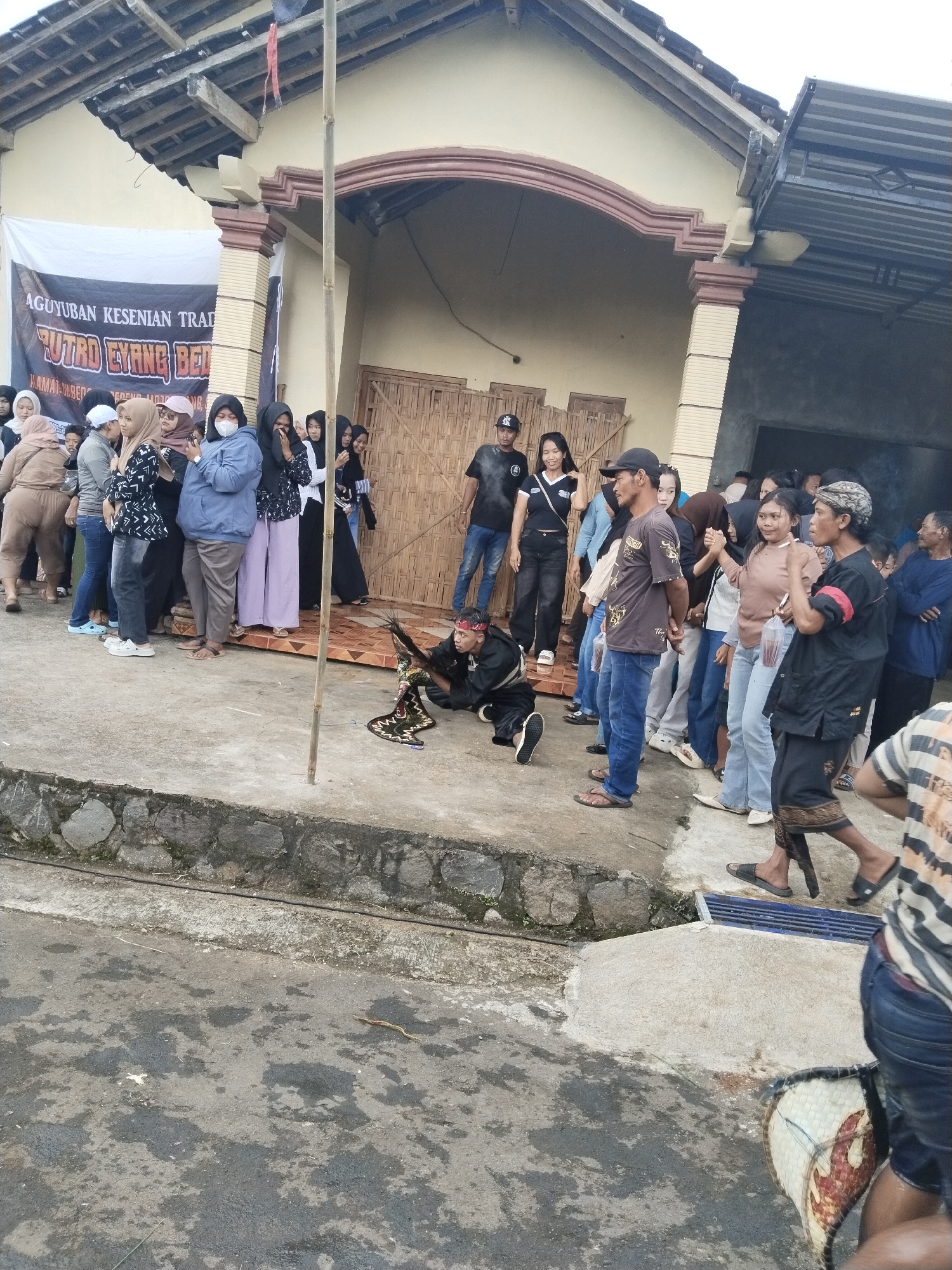 The scene depicts an outdoor gathering in Pendem, Indonesia. A group of people are congregated in front of a building with light-colored walls and a dark roof. Some individuals are standing in a line, while others are observing. A person in a black outfit is kneeling on the ground, holding what appears to be a decorative object. The ground is concrete with some visible cracks and stains. A low stone wall separates the foreground from the area where the people are standing. A banner with text is partially visible on the left side of the building.