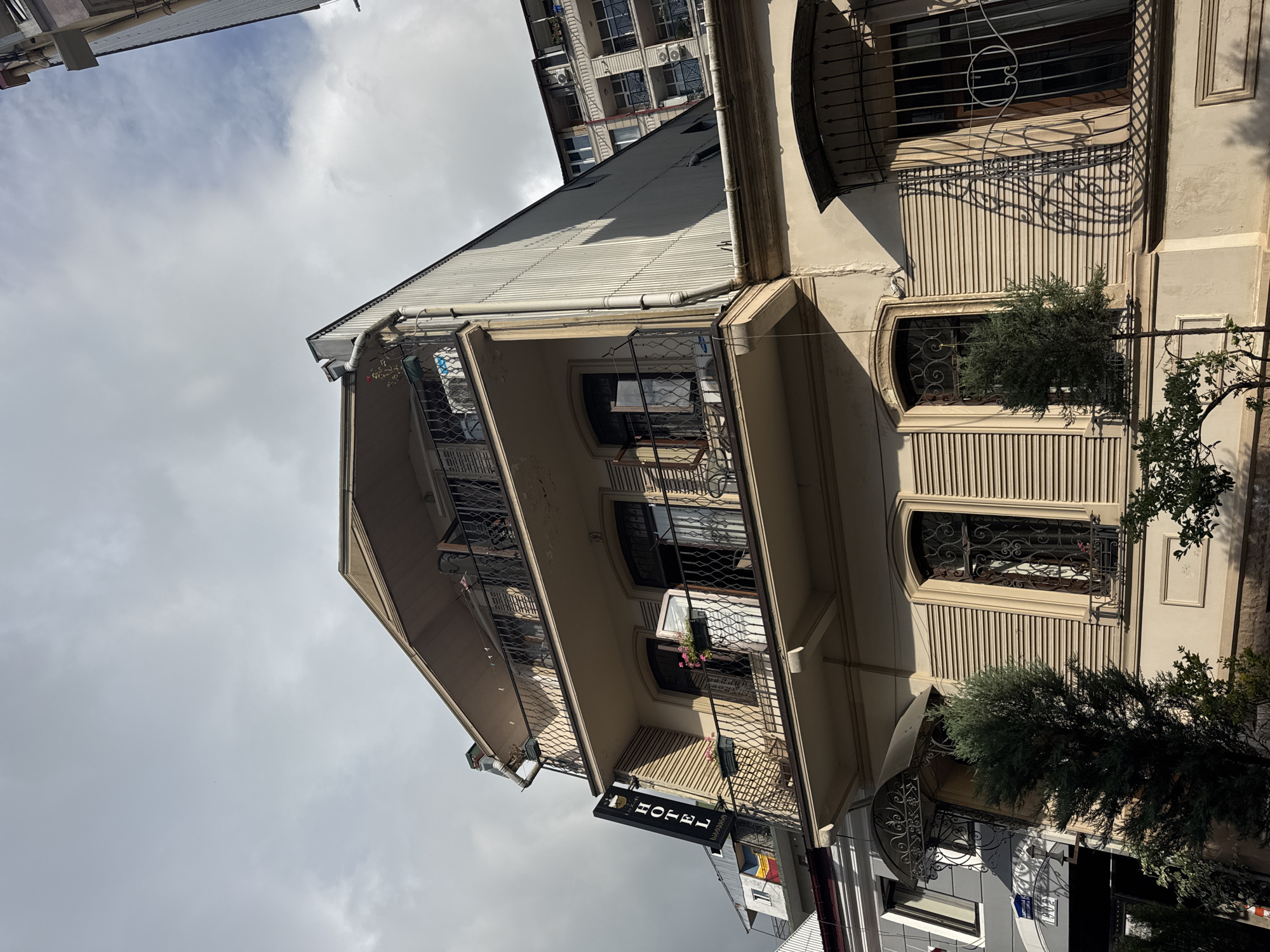 The image shows a street-level view of a building with a facade of light-colored plaster. The building has multiple stories with balconies on the upper levels, protected by decorative metal railings. Several windows are visible, some with similar metal grilles, and one window on the second floor has a small planter with pink flowers. A dark-colored sign hanging from the building reads "HOTEL" in white, sans-serif lettering. The architectural style appears to be a mix of older and more modern elements. To the left of the building, another structure, possibly an apartment building, rises higher, with visible air conditioning units on its balconies. Trees are planted in front of the building, one is a tall, slender evergreen, and another is a more leafy deciduous tree partially obscuring the lower part of the image. The sky above is overcast with clouds, suggesting a cool or moderate temperature. The photograph is taken from a low angle, looking upwards, and the composition is tilted. The overall scene is an urban streetscape in Batumi, Georgia, with a focus on a hotel. No people are visible, and the time of day is indeterminate due to the overcast sky.