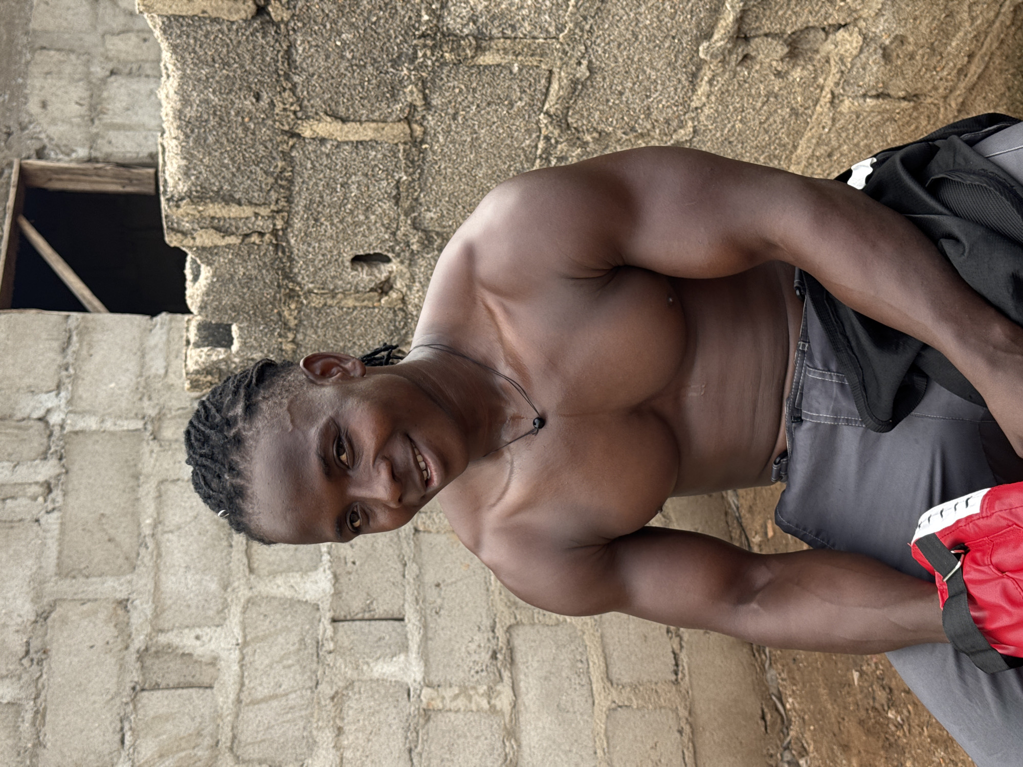 A man with braided hair and a muscular physique is sitting in front of a brick wall. He is shirtless and wearing grey pants. A red and black bag is resting on his lap. The environment appears to be an outdoor setting, possibly in Akwanga, Nigeria, with a rough brick wall as the backdrop. The man is smiling at the camera.