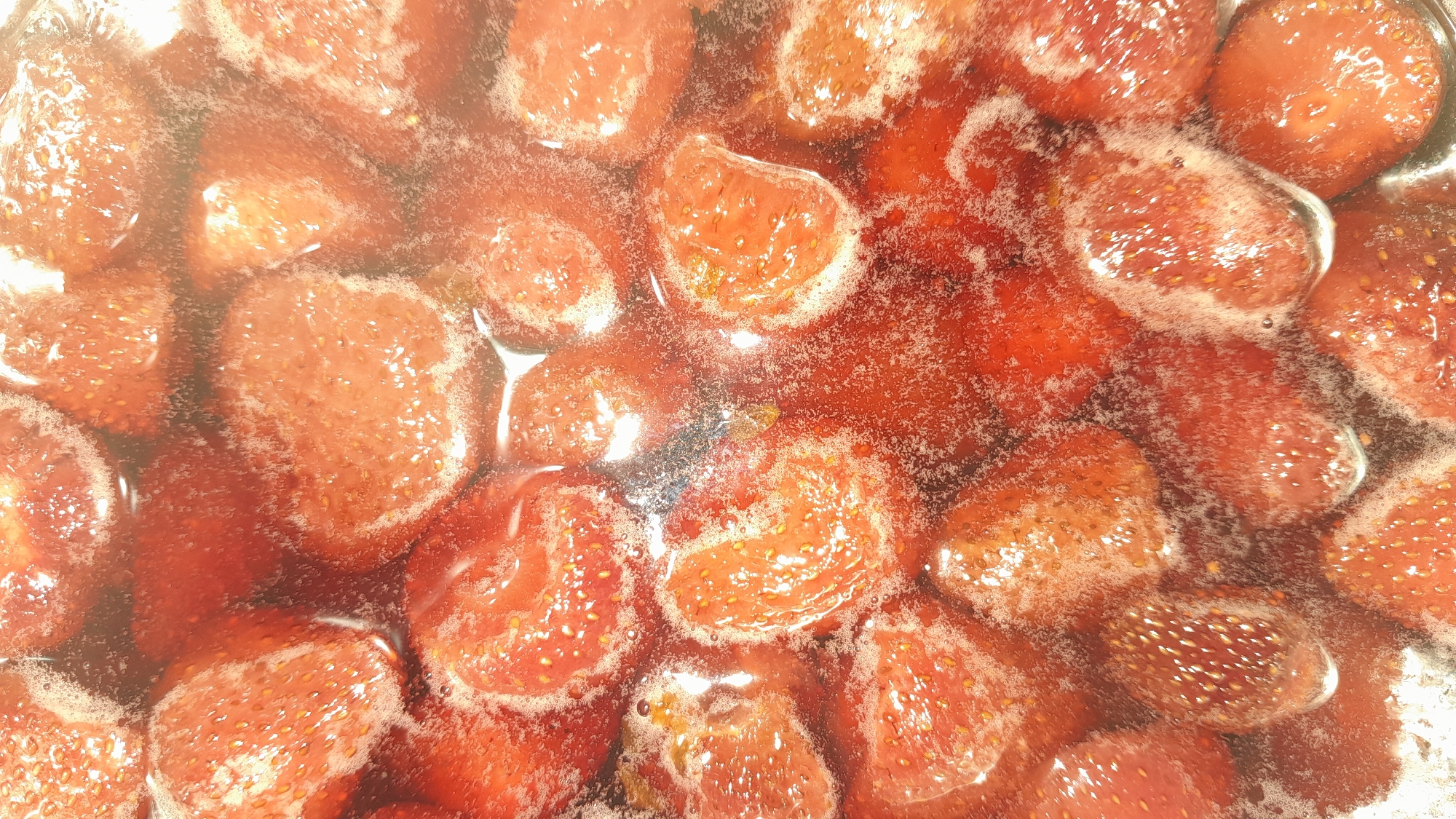 The image is a close-up, overhead shot of strawberries simmering in a liquid, likely being cooked into jam or preserves. The strawberries are whole or halved, and appear softened and slightly shrunken. They are submerged in a reddish, syrupy liquid that is bubbling and frothing around the fruit. The lighting is bright, creating reflections and highlights on the surfaces of the strawberries and the liquid, giving the scene a glossy appearance. The overall impression is one of a food preparation in progress. The image does not provide any information about a specific location in a city.