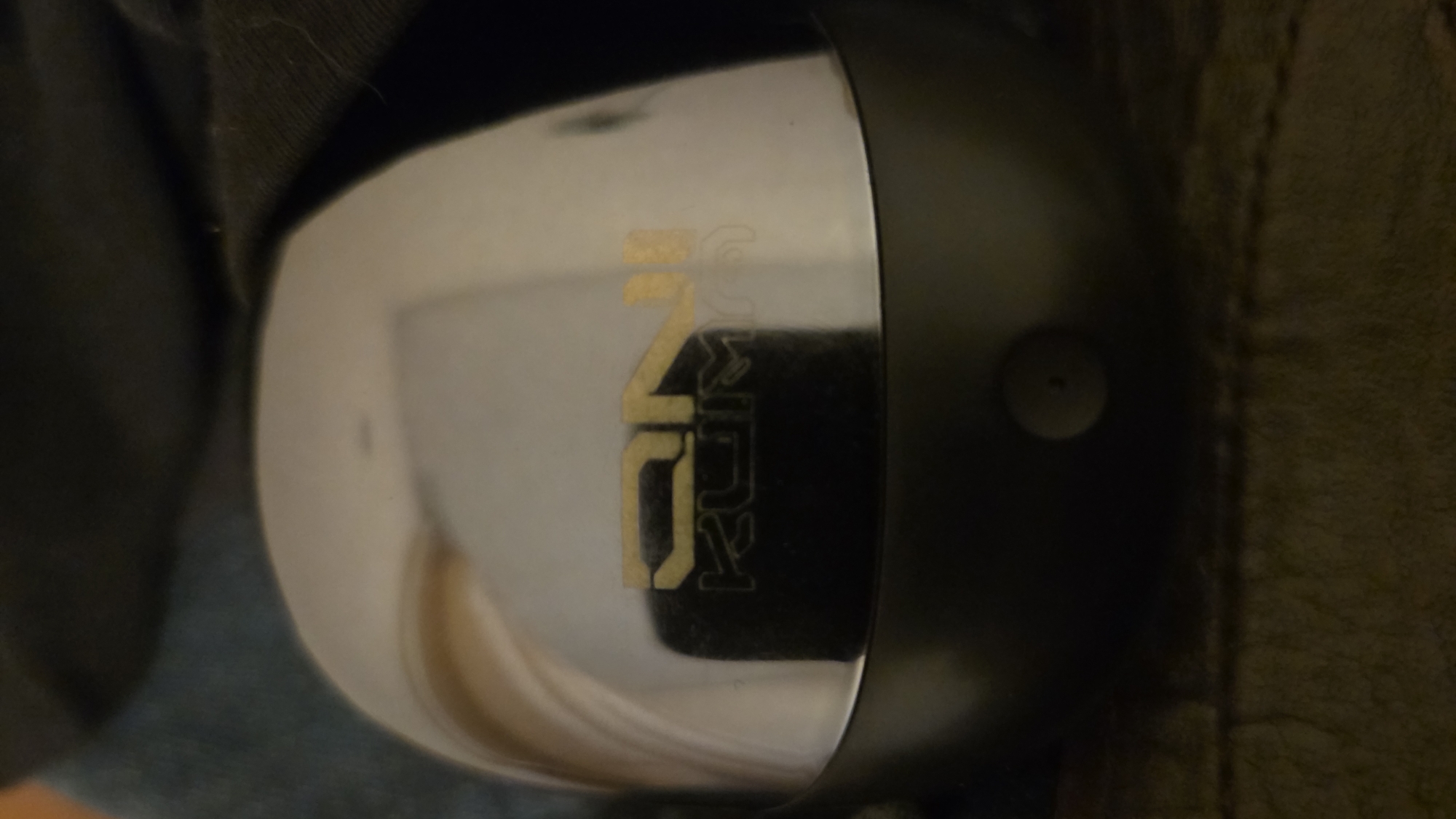The image shows a close-up, overhead view of a chrome-plated, rounded object, possibly a lid or a cap, resting on a dark, textured surface. A gold-colored logo is visible on the object, with the letters "DN" at the top and "KUM" below. There is a small, circular button or sensor near the bottom edge of the object. The background is out of focus, showing dark fabric and some hints of blue material, possibly clothing. The scene appears to be indoors, with no specific geographical markers to indicate a location within Karaganda, Kazakhstan. The lighting is dim, creating reflections on the chrome surface.