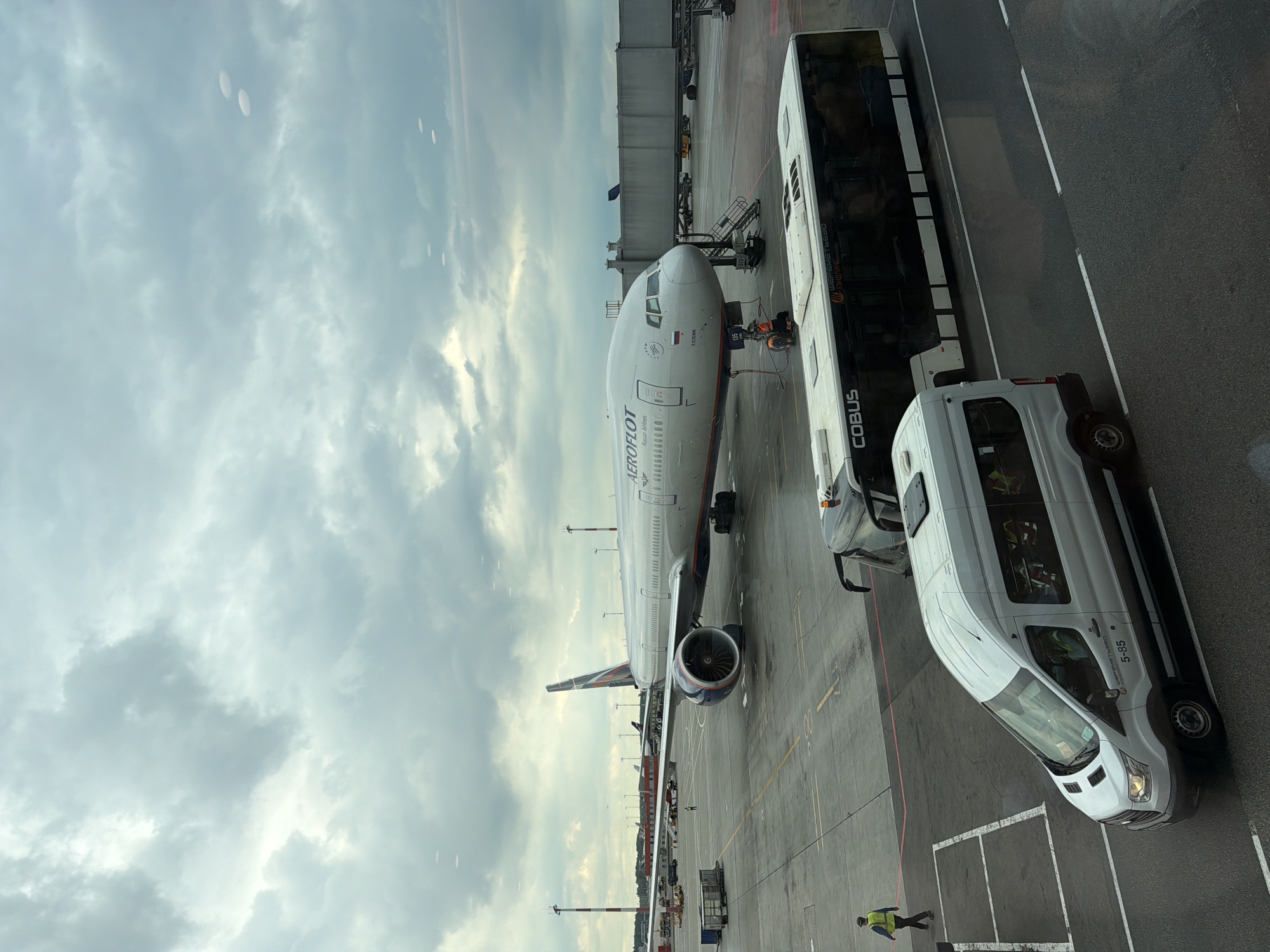 An Aeroflot passenger jet is parked at an airport gate under a cloudy sky. The aircraft, a white Boeing 777 with the Aeroflot logo and "Russian Airlines" text on its fuselage, is positioned on the tarmac. A jet bridge connects to the aircraft. In the foreground, two buses are present; one is a white passenger van with tinted windows, and the other is a larger, dark-windowed bus with "COBUS" branding. A person in a high-visibility vest is visible walking near the white van. The ground is wet, suggesting recent rain. The scene appears to be at Sheremetyevo International Airport (SVO) in Khimki, Russia.