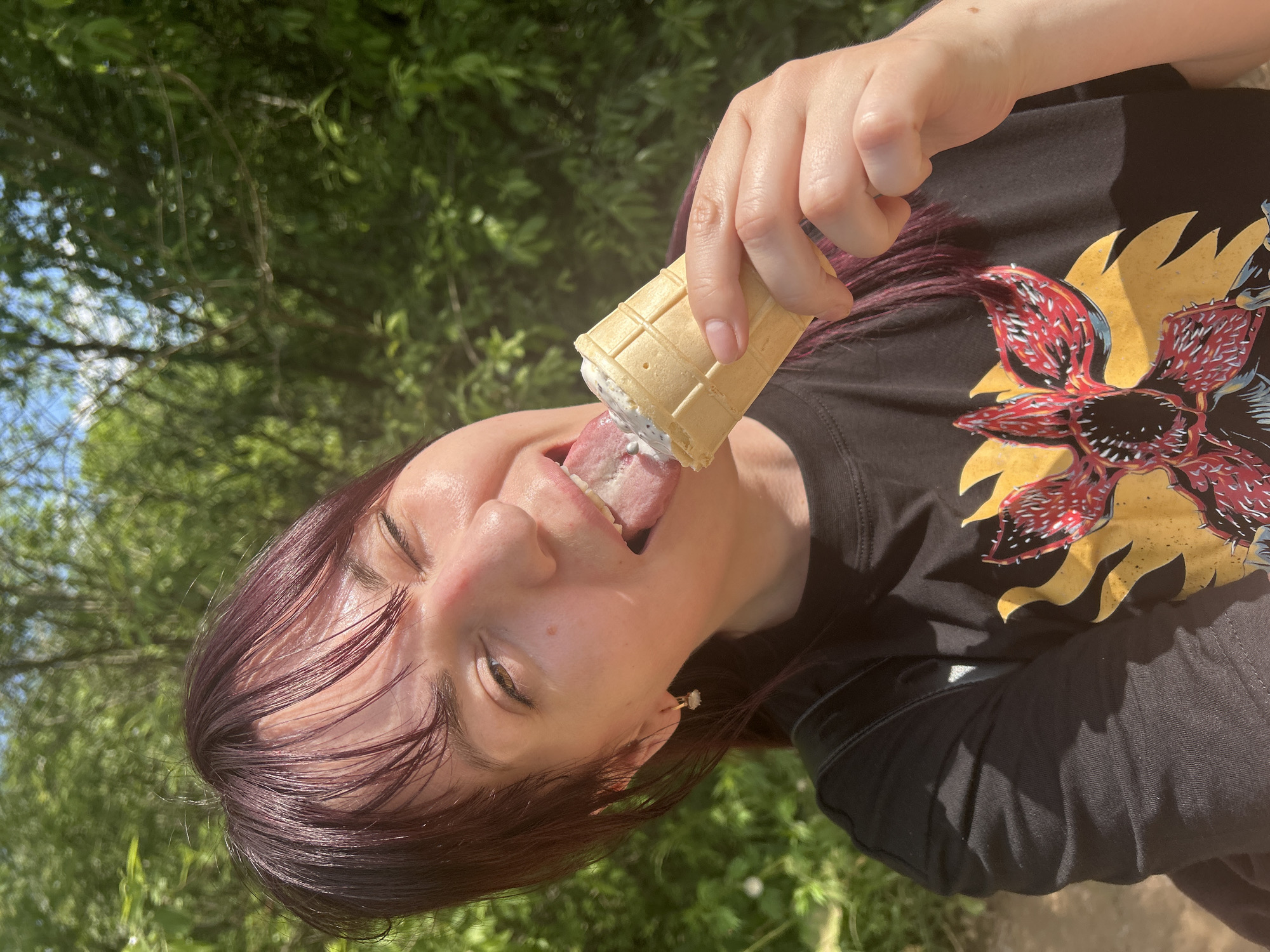 A woman with dark purple hair is pictured outdoors, in what appears to be a wooded area with green foliage and a bright sky. She is holding an ice cream cone to her mouth and sticking her tongue out to lick it. The woman is wearing a black t-shirt with a graphic design of a stylized red and yellow flower-like creature. The image is taken from the chest up, with the woman's face prominently featured. The lighting suggests it is daytime, and the overall atmosphere is casual and summery. The location is stated to be in the city of Фоки, Russia.