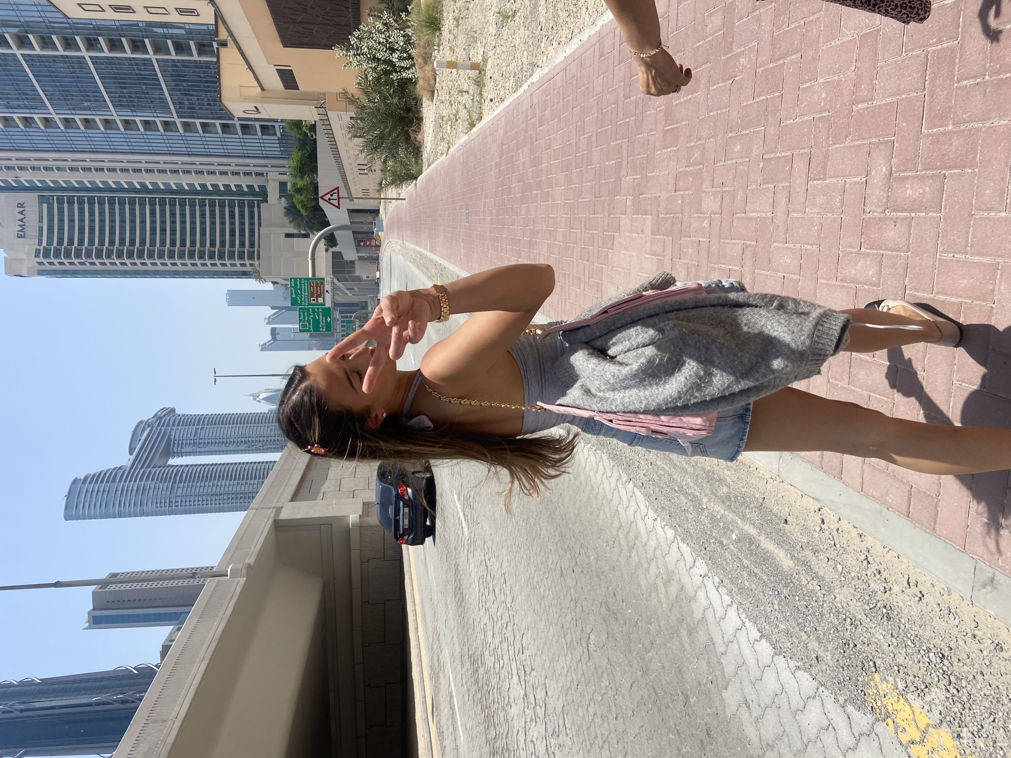 A horizontal shot captures a woman standing on a brick-paved sidewalk, posing with a peace sign. She wears a gray top, a denim skirt, and a gray sweater draped over her shoulder. A pink purse hangs from her shoulder. To the left, a black car is parked.
The background features the urban landscape of Dubai, United Arab Emirates. Tall buildings, including one with the "EMAAR" logo, dominate the skyline. Street signs are visible, indicating directions to various locations like Sheikh Zayed Road and Burj Khalifa/Dubai Mall. The clear sky suggests a sunny day. In the top right corner, a hand with a bracelet is visible.