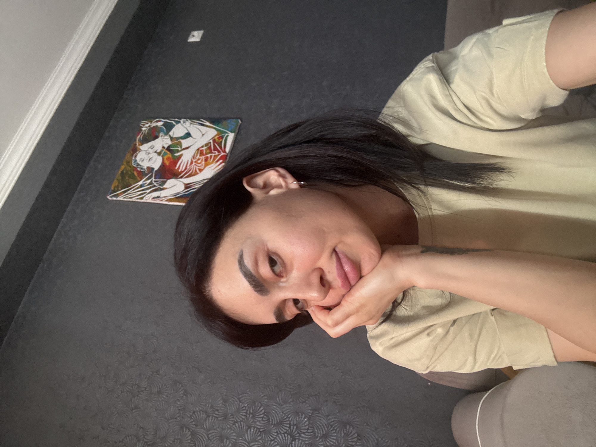 A medium shot shows a woman in a tan shirt with her face resting in her hand, looking at the camera. She has dark hair, prominent eyebrows, and a piercing in her ear. A piece of art hangs on a dark gray textured wall behind her. The location appears to be indoors, in Tbilisi, Georgia.