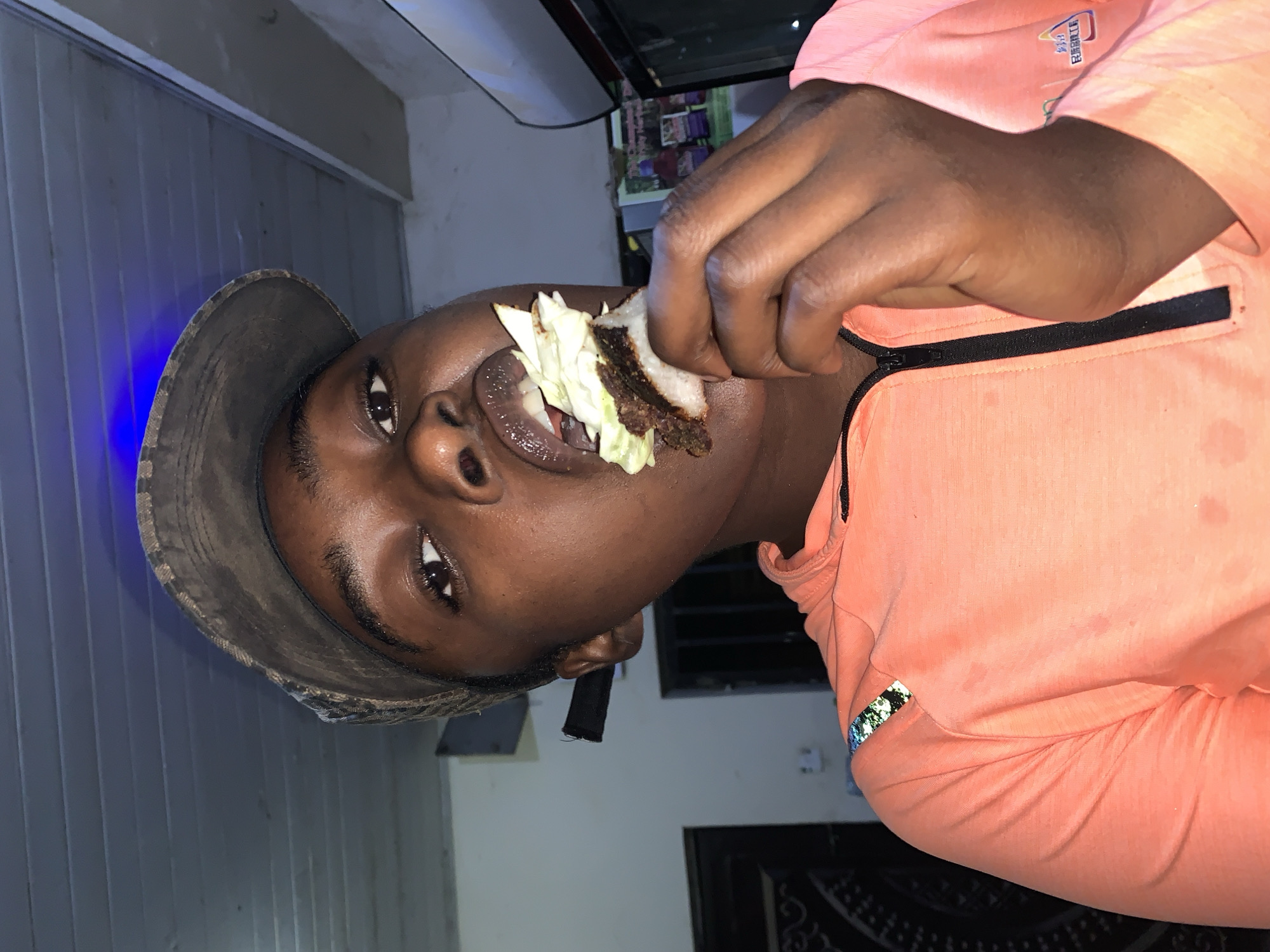 A person is eating a sandwich or burger-like food item with lettuce. The person is wearing a hat and an orange shirt with a black zipper. The setting appears to be indoors or near a building, possibly in the city of New Stadium, Nigeria based on the request. There is a window with blinds in the background.