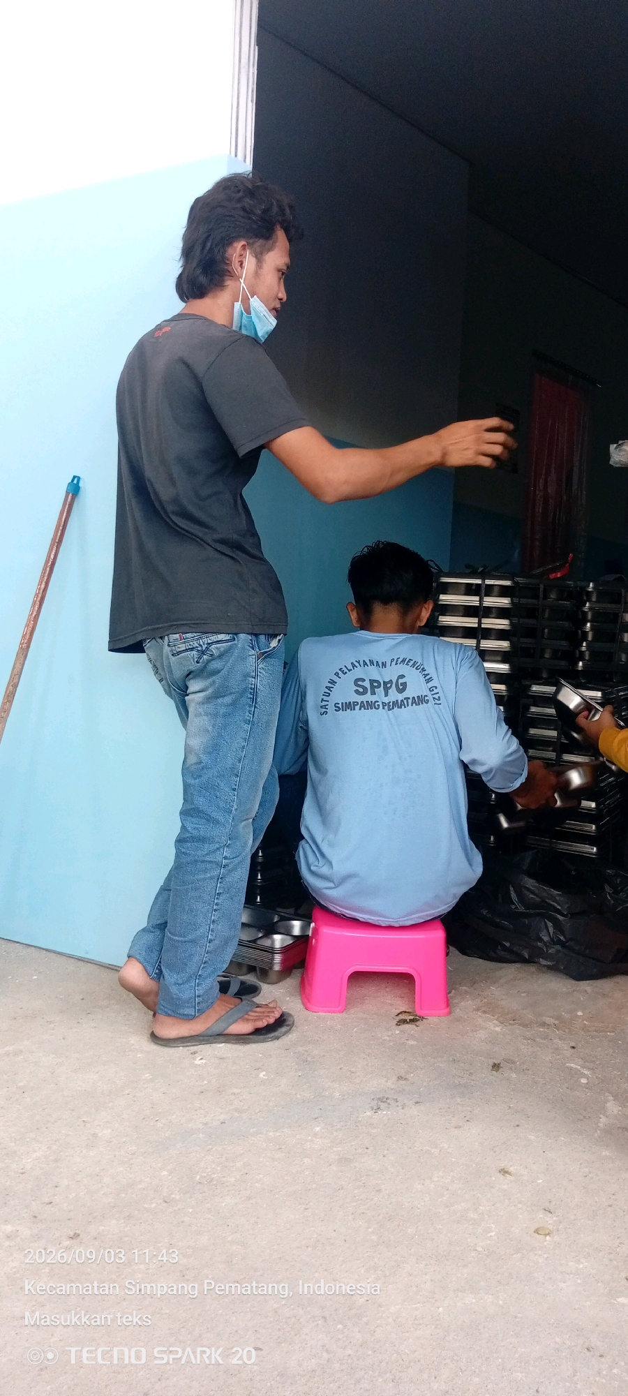Two men are visible in an outdoor urban setting. The man on the left is standing and wearing a dark t-shirt, blue jeans, and sandals. He has a light blue face mask pulled down to his chin. He is holding a dark object in his right hand, reaching towards the right. The man on the right is seated on a pink stool, facing away from the camera. He is wearing a light blue long-sleeved shirt with text on the back and dark pants. He is looking down at his hands, which are near a metal tray. In the background, there are stacks of metal trays and a large black plastic bag. The ground is a light-colored concrete surface. Text overlay indicates the date and time as "2026/09/03 11:43" and the location as "Kecamatan Simpang Pematang, Indonesia". The text "Masukkan teks" and "TECNO SPARK 20" are also present.