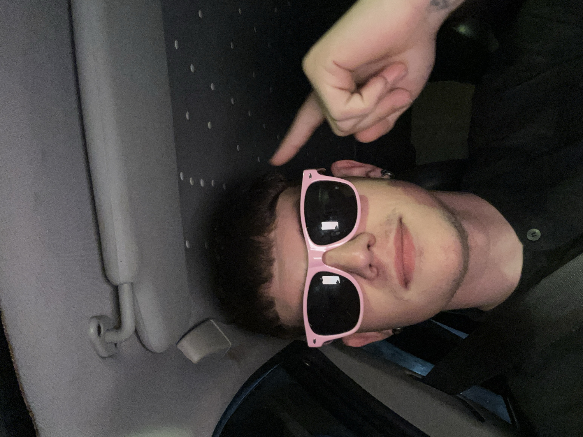 A person with short, dark, textured hair and light skin is taking a selfie inside a vehicle. They are wearing pink Wayfarer-style sunglasses, a black collared shirt with a button placket, and black hoop earrings. Their right index finger is raised and pointing towards their forehead, near the sunglasses. The person is looking directly at the camera and has a slight smile. The interior of the car is visible, including a grey sun visor with a hinge mechanism on the left, and a dark headliner with small, circular lights. The seatbelt is visible across the person's chest. The lighting suggests it is nighttime or a dimly lit environment.