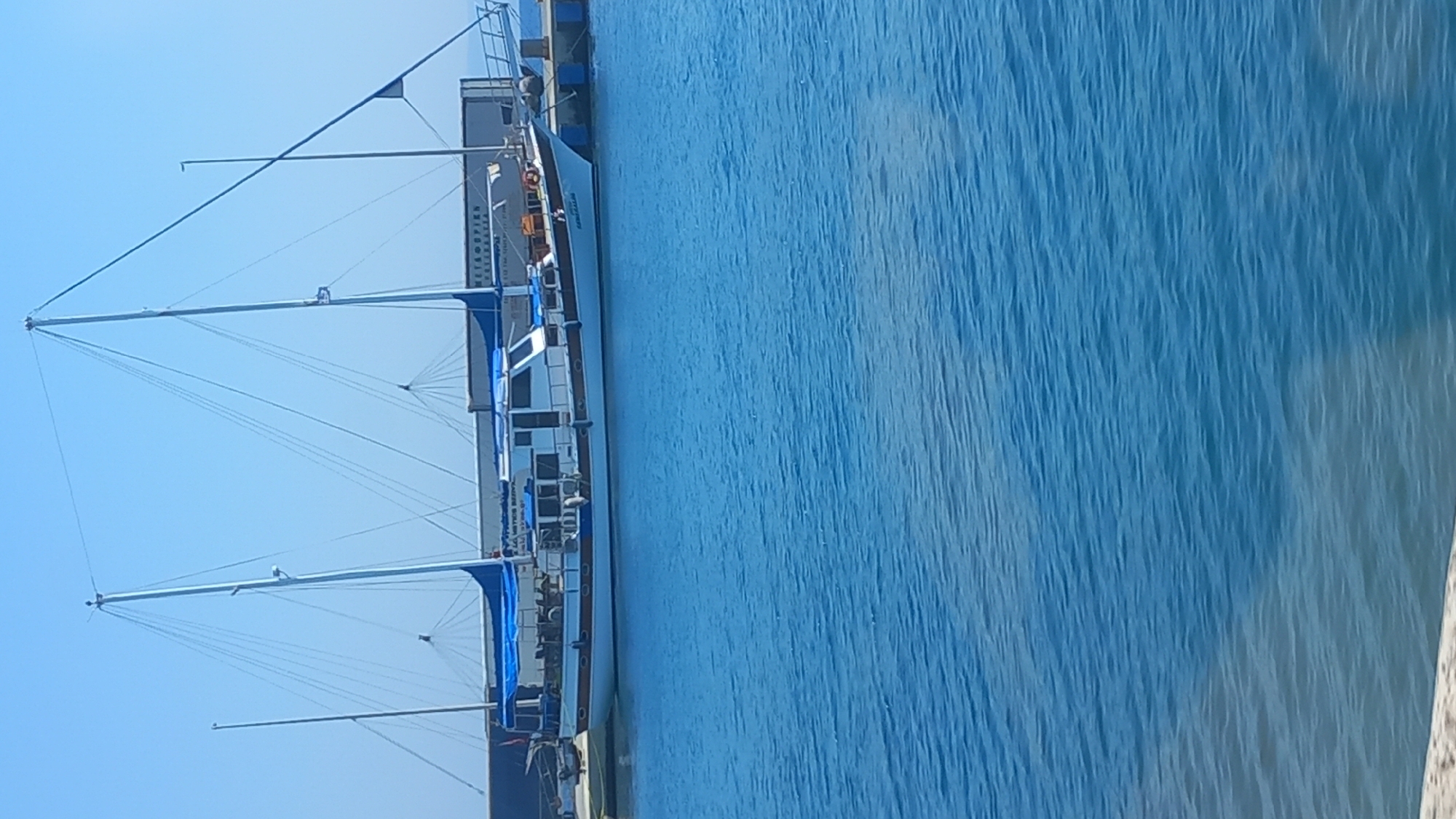 A sailboat with blue sails and a white hull is docked in the city of Kos, Greece. The sailboat is moored at a pier with a blue border. In the background, there is a clear blue sky and a glimpse of a large cargo container. The water is a vibrant blue and appears calm. The image captures a sunny day in the port of Kos.
