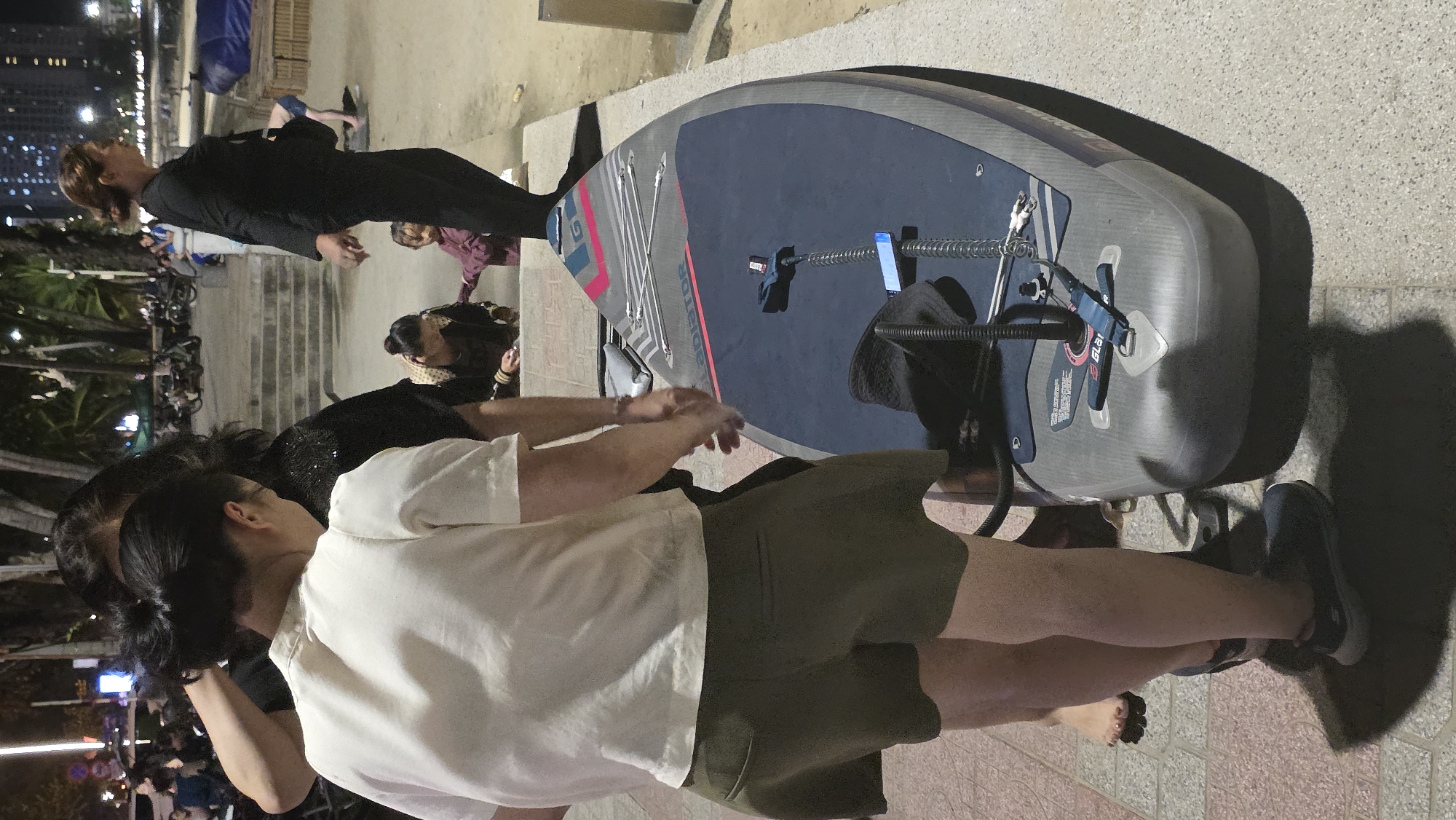 The image depicts a group of people next to a paddleboard, which is lying on the ground. The paddleboard is mostly gray with blue and pink accents. One person is standing near the paddleboard, wearing a white shirt and khaki shorts. There are people visible in the background, some standing and some sitting. The setting appears to be an outdoor area, likely near a beach, as evidenced by the presence of palm trees and people in casual attire. A city backdrop with multiple buildings is visible in the distance, suggesting this is the Nha Trang Ward in Vietnam.