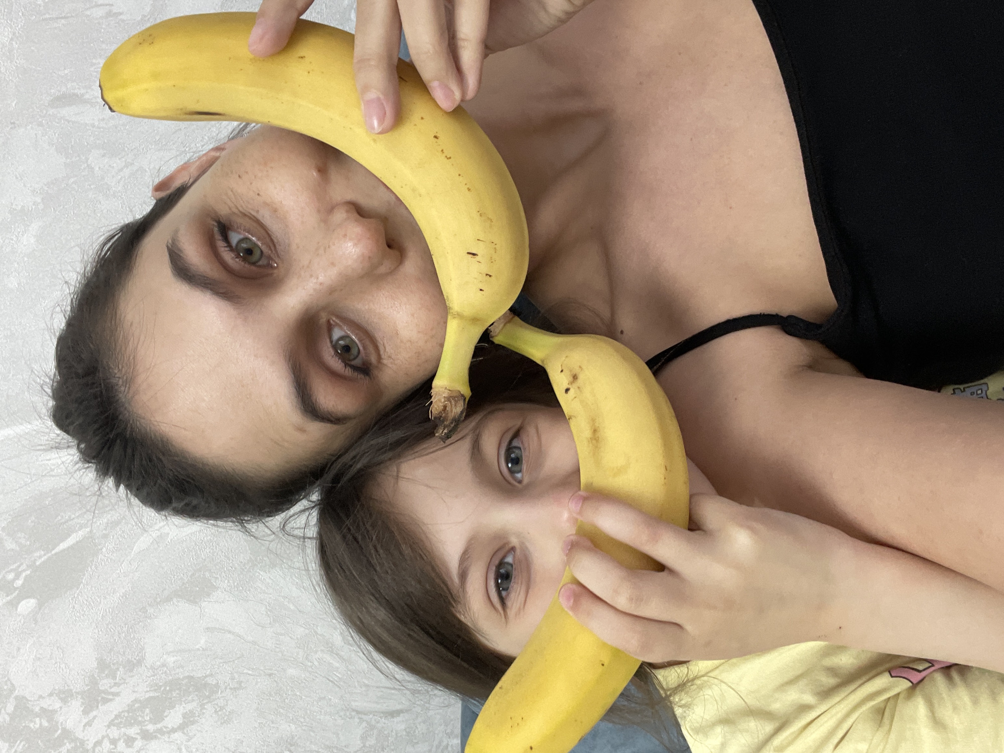 This image, captured in Izhevsk, Russia, features two individuals – an adult and a child – in a playful pose, viewed from a top-down perspective. The image is oriented vertically, with the adult positioned towards the top and the child below.
The adult, who has dark hair, green-hazel eyes, and noticeable freckles, wears a black top with visible straps. She holds a ripe yellow banana above her face, covering parts of her forehead and hair, with her manicured fingers visible near the banana's upper curve. Her expression is engaging, looking directly at the camera with wide, curious eyes.
Below the adult, a child with brown hair and light blue eyes similarly holds a ripe yellow banana across her face. Her small hands grasp the banana on either side, framing her eyes as she also looks directly at the camera with an inquisitive and joyful gaze. She appears to be wearing a yellow garment.
The background consists of a plain, light grey or off-white textured surface, suggesting an indoor setting, possibly a wall or bedding. The overall scene conveys a sense of lighthearted fun and familial intimacy between the two subjects. There is no visible text in the image.