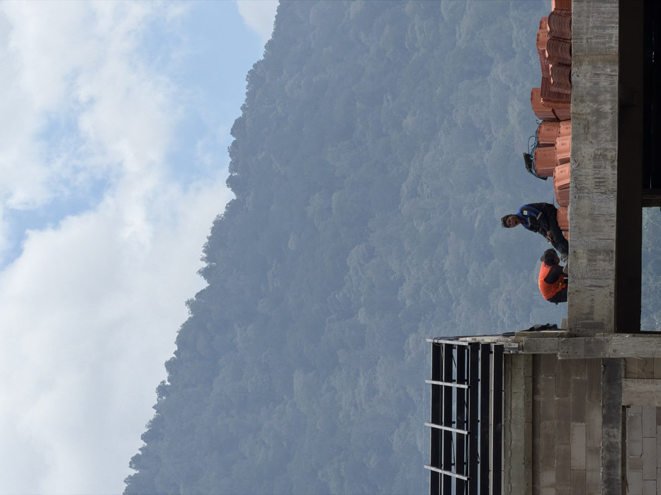 Two construction workers are working on the roof of a partially constructed building with a mountain in the background. The man standing is wearing a blue shirt and dark pants, and the man kneeling is wearing a red shirt. The building appears to be made of cinder blocks and has a stack of roof tiles on top of it. The sky is visible behind the mountain.