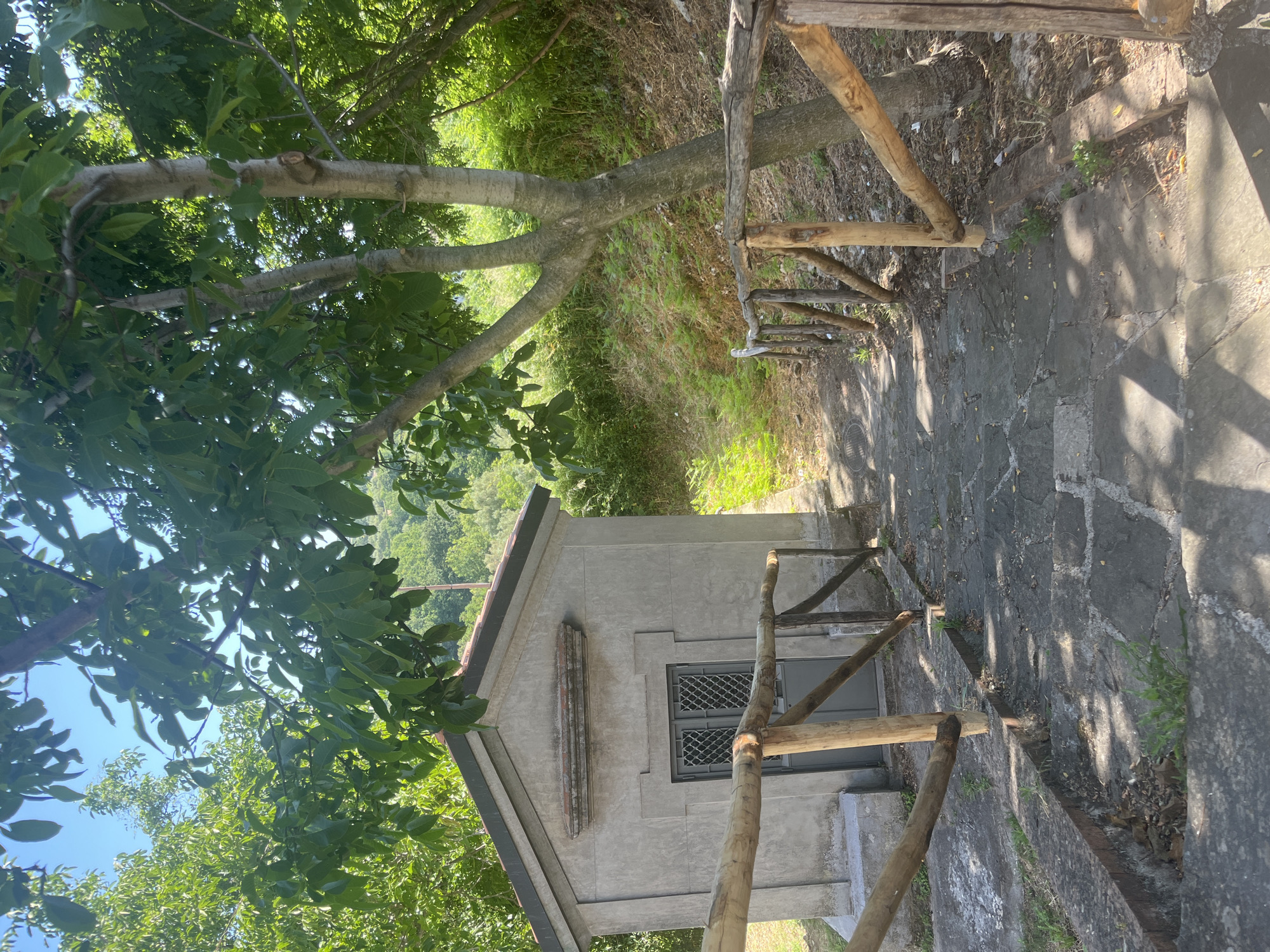 The image shows a small, grey, rectangular building with a gabled roof, possibly a chapel or mausoleum, situated on a hillside in a verdant, wooded area. A stone pathway, bordered by a rustic wooden fence, leads up to the building. The pathway is made of irregular flagstones and is partially covered in shadows from the surrounding trees. The building has a grey door with a metal grate. The environment suggests a rural or park-like setting, with dense green foliage and a clear blue sky visible. The image is taken from a slightly elevated perspective, looking down the pathway towards the building. The overall scene is one of quiet solitude and natural beauty.