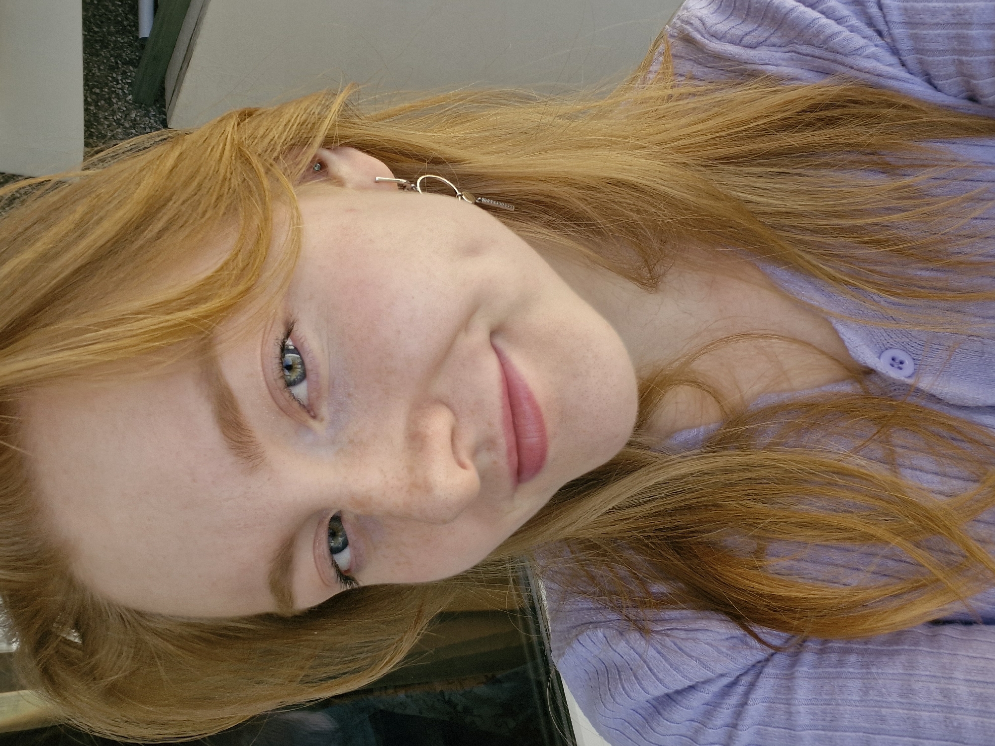 A young woman with long, reddish-blonde hair and light skin is seen from a close-up, slightly angled perspective. Her eyes are light blue-green, and she has freckles on her face. She is wearing a lavender-colored, buttoned sweater. An earring is visible in her left ear. The background is indistinct but appears to include a light-colored wall or surface. The image does not contain any information related to Yekaterinburg, Russia.