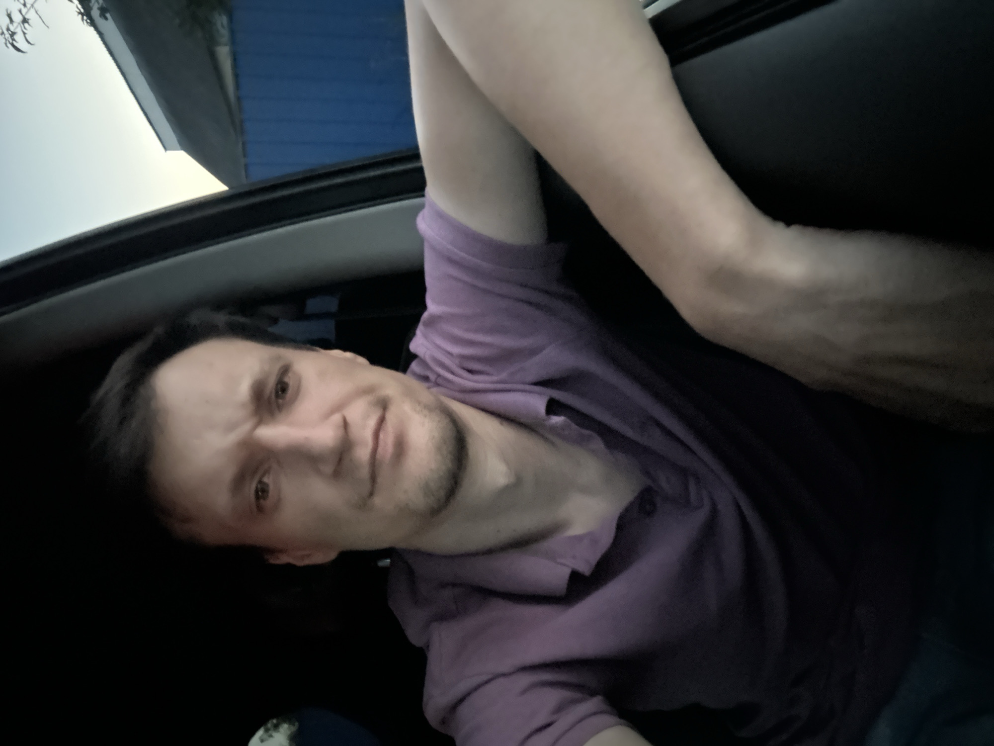 A man is in the driver's seat of a car, looking at the camera. He is wearing a purple polo shirt. His left arm is resting on the window frame, and his right arm is out of view. Outside the car, a blue fence and a roof are visible. The sky is a pale blue, indicating it is either dawn or dusk. The scene appears to be in a residential area. The image was taken on July 1, 2023, at 8:44 PM, in Shatava, Ukraine.
