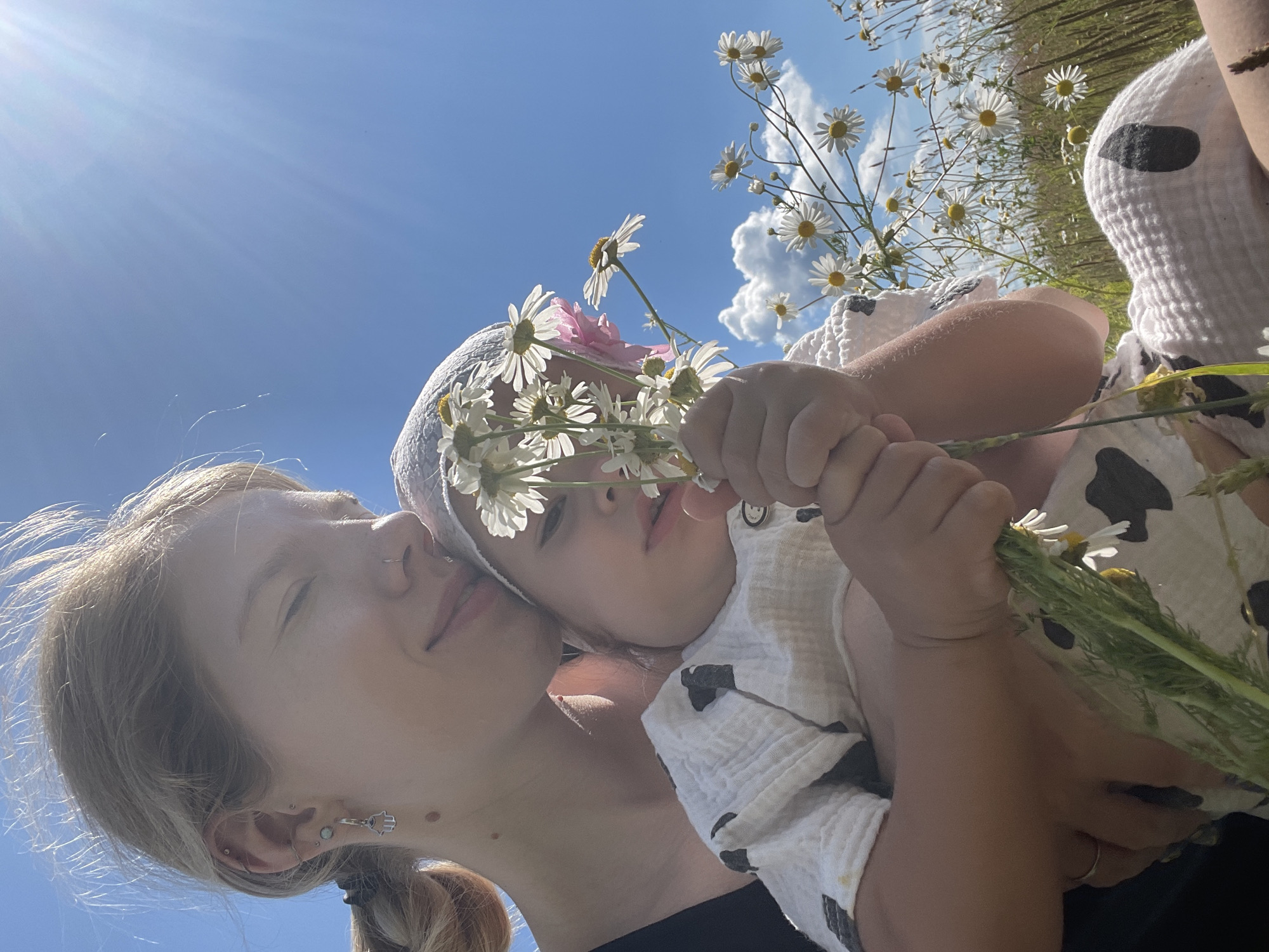An outdoor, low-angle, medium shot captures a fair-skinned young woman holding a baby in a field of daisies under a bright blue sky. The woman, with blonde hair pulled back, is wearing a black top and has several earrings and a nose ring. She is smiling with her eyes closed as she embraces the baby. The baby, wearing a white patterned outfit and a white hat, is holding a small bouquet of daisies. The field is filled with numerous white daisies and green grass, with scattered clouds visible in the sky. The sun is shining brightly, creating lens flare in the upper right portion of the image. The location is not explicitly stated to be in the city of Сям-Можга, Russia, but rather an outdoor natural setting.