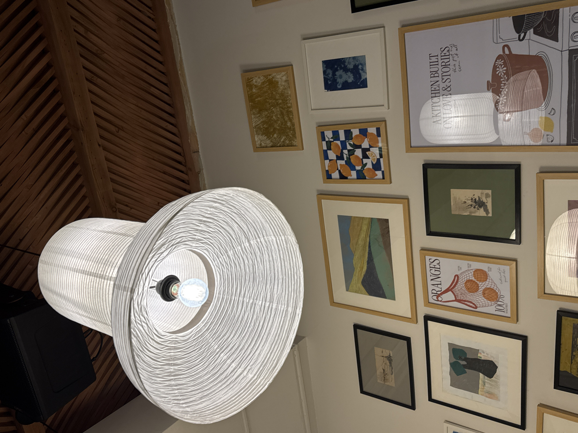 A low-angle indoor shot depicts a large, white, cylindrical pendant lamp hanging from a ceiling with wooden paneling in Yerevan, Armenia. The lamp has a spiral design and a visible light bulb within. The wall behind the lamp is decorated with various framed artworks, including images of lemons, abstract designs, and kitchenware. The lighting in the room is dim.