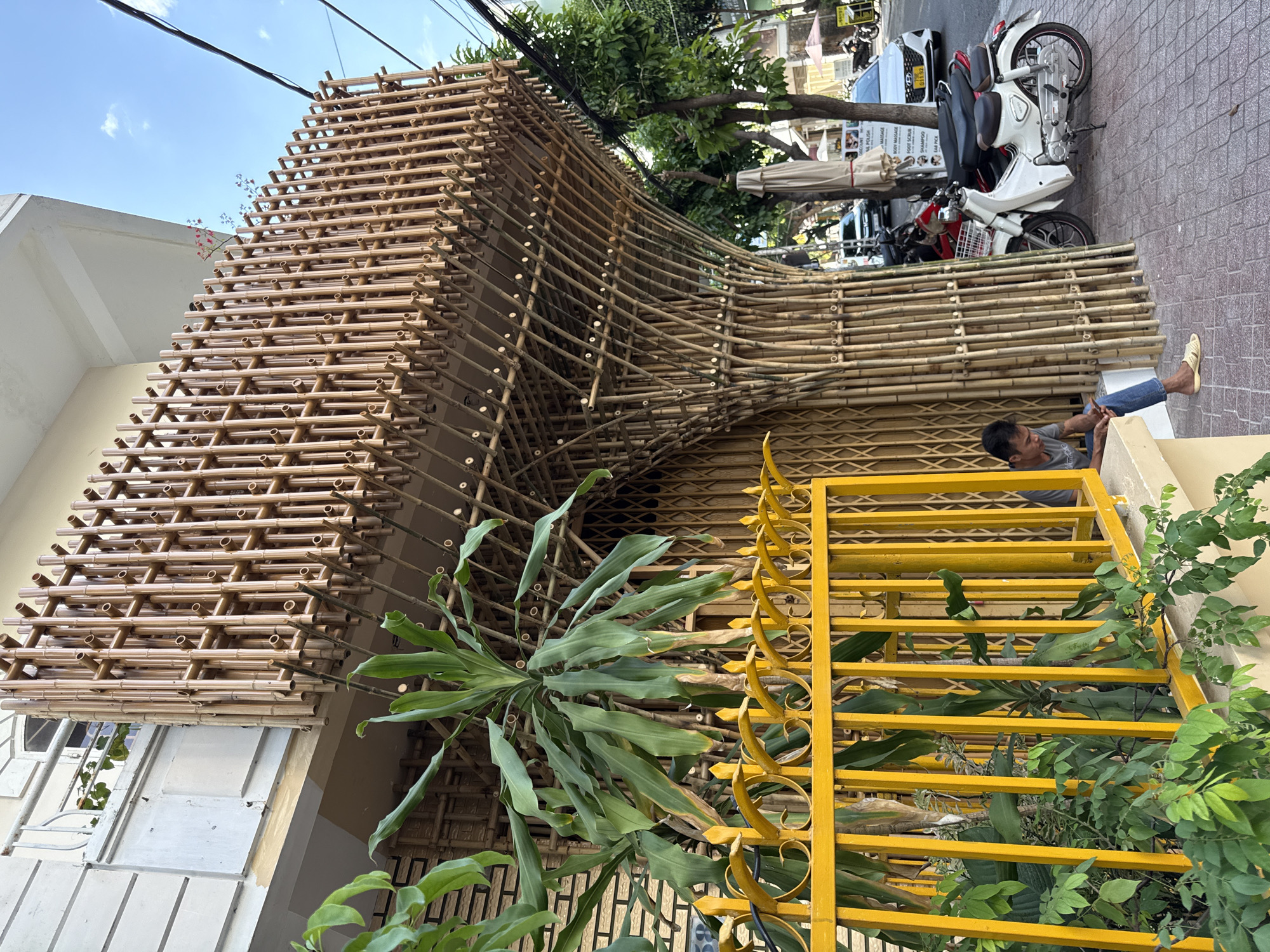 In Nha Trang, Vietnam, a man sits on a low wall to the left of a building with an elaborate bamboo facade. The facade is constructed from numerous vertical bamboo poles, creating a visually complex and organic architectural feature. The bamboo structure extends from the ground level and curves upward to form an awning or balcony overhanging the sidewalk. A bright yellow metal fence with a decorative scroll pattern is in the foreground, partially obscuring a lush green plant. Behind the fence and to the left of the bamboo structure is a closed, accordion-style metal gate. Several motorcycles are parked on the sidewalk to the right of the bamboo facade. The street is paved with hexagonal tiles, and in the background, cars, trees, and other buildings are visible under a bright blue sky with scattered clouds. The man, wearing a gray t-shirt and blue jeans, is looking down at his mobile phone.