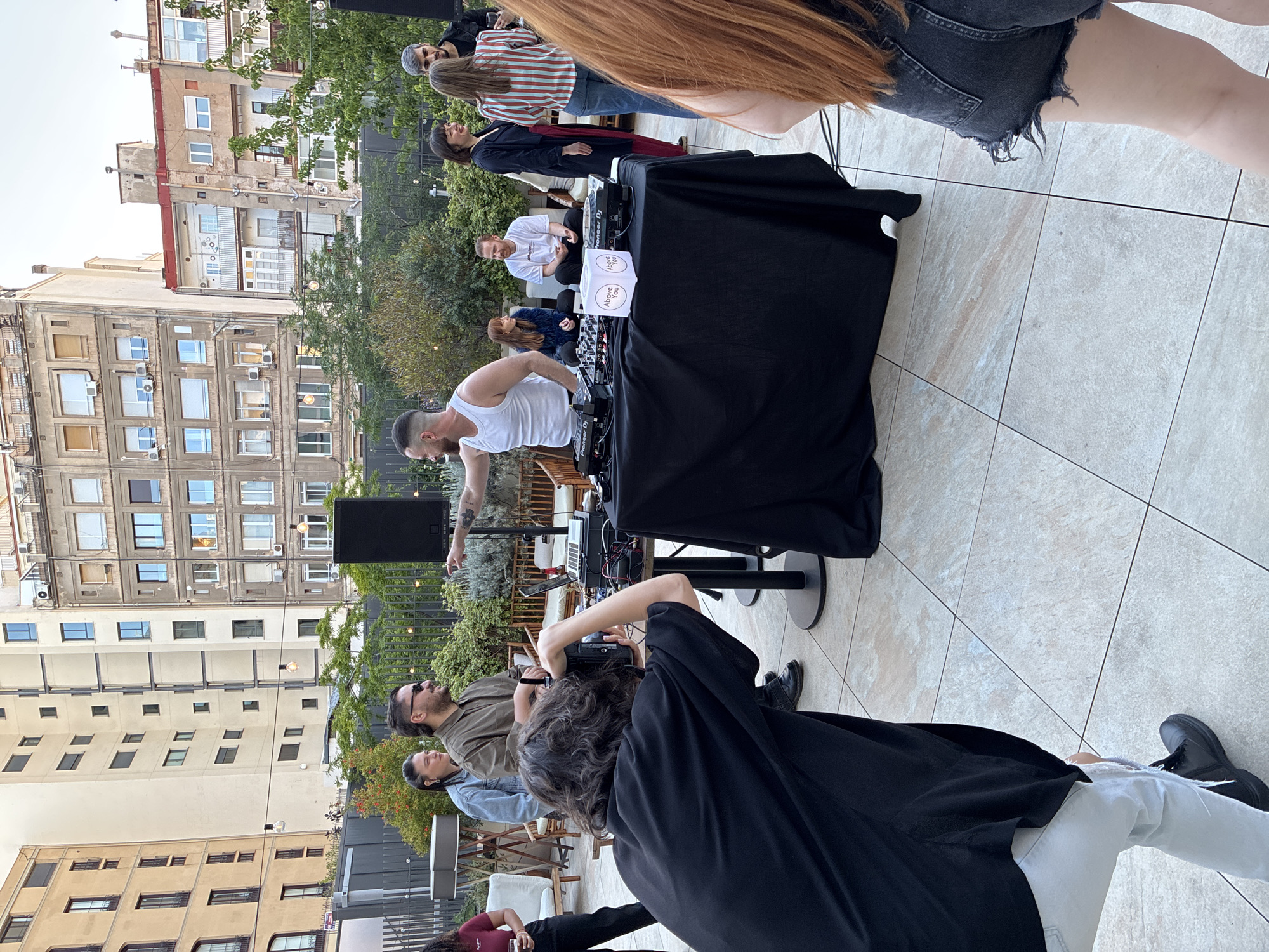 The image depicts an outdoor gathering, possibly a small event, on a rooftop terrace. Several people are present, some of whom appear to be performing or operating sound equipment. A DJ in a white tank top stands at a table covered with a black cloth, with audio equipment set up on top. There are two cubes on the table labeled "Above You." Another person in a white t-shirt is sitting cross legged behind the table.
The environment includes greenery, hinting at a garden-like setting, with buildings of varying heights and architectural styles forming the backdrop. The buildings resemble the urban landscape found in cities like Barcelona, Spain. The rooftop has light-colored tiles. The sky is overcast. The photograph is taken at an angle, with people facing different directions. The overall scene suggests a social gathering, potentially with music or entertainment.